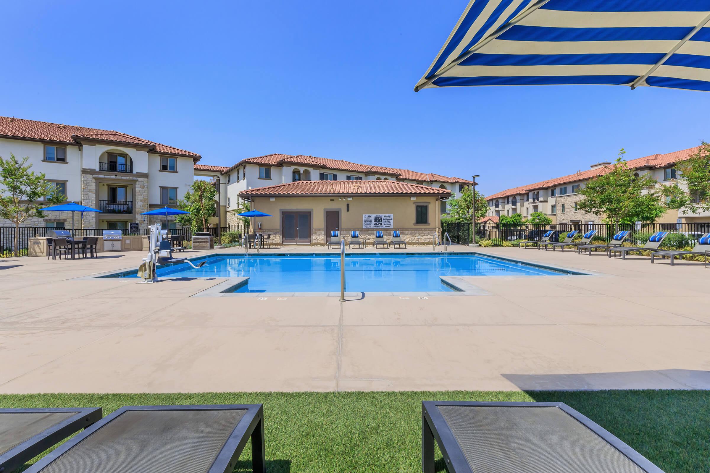 A clear blue swimming pool surrounded by lounge chairs and umbrellas, set in a residential complex. The pool area features several trees and landscaped areas, with modern buildings in the background under a sunny sky.