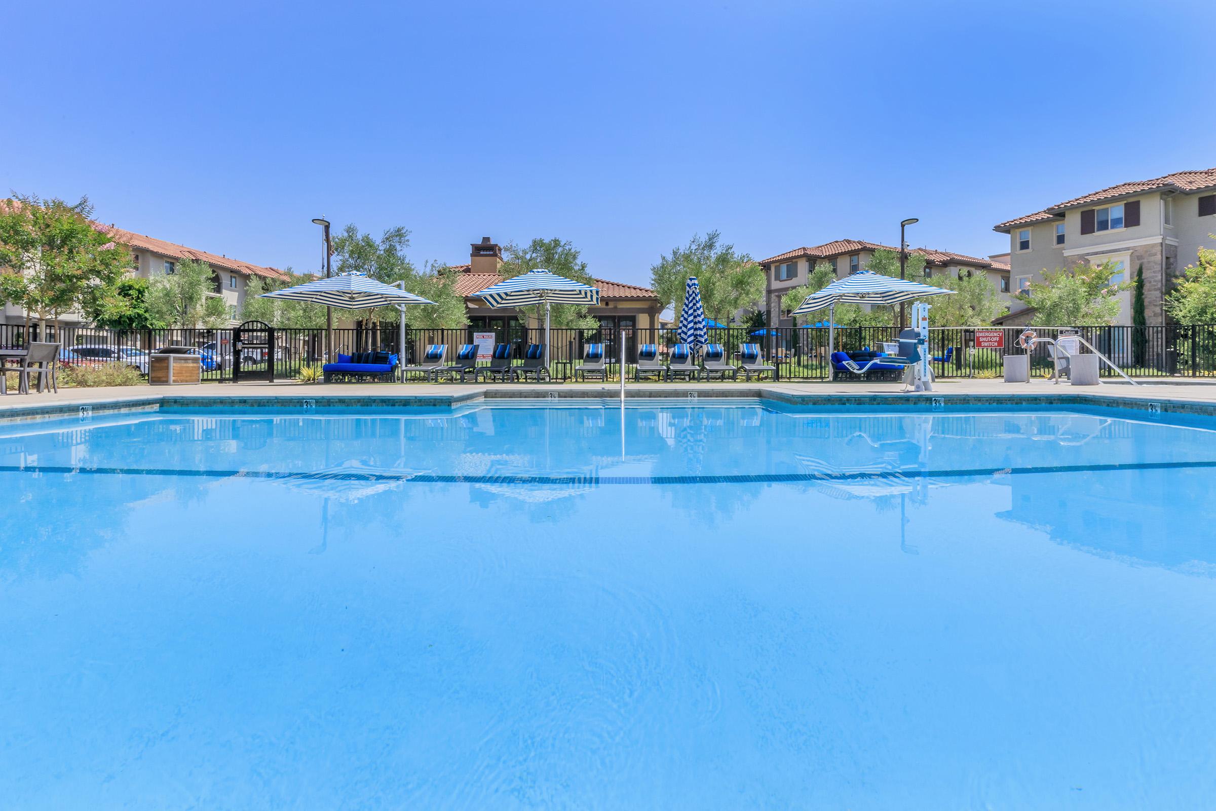 A clear swimming pool surrounded by lounge chairs and umbrellas, located in a residential area. The area features well-maintained greenery and several buildings in the background under a bright blue sky.