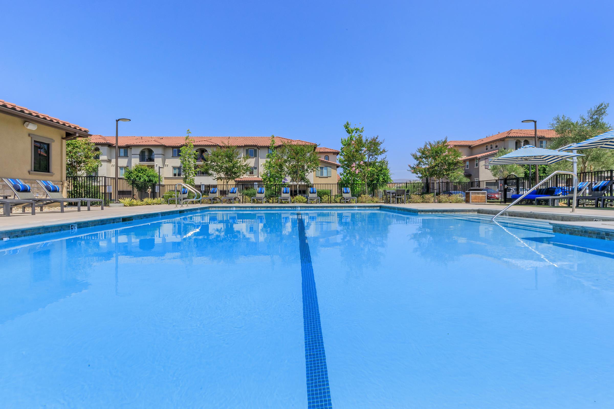 A clear, blue swimming pool surrounded by lounge chairs and lush greenery. The pool area features a bright, sunny sky and residential buildings in the background, creating a relaxing outdoor atmosphere.