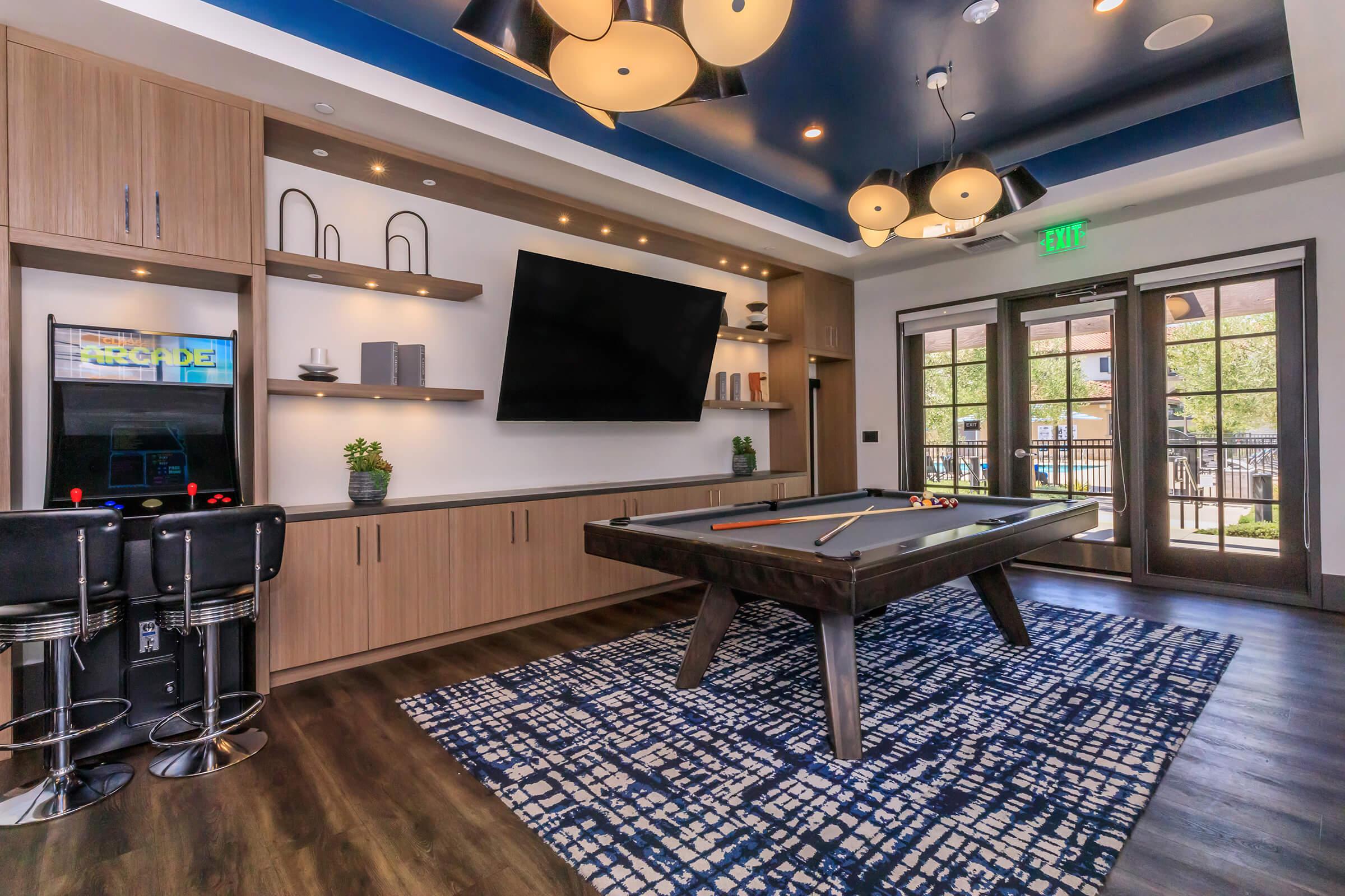 A modern recreational room featuring a billiard table, stylish bar stools, a large flat-screen TV mounted on the wall, and contemporary lighting fixtures. Large windows allow natural light, and a patterned area rug adds color to the wooden floor.