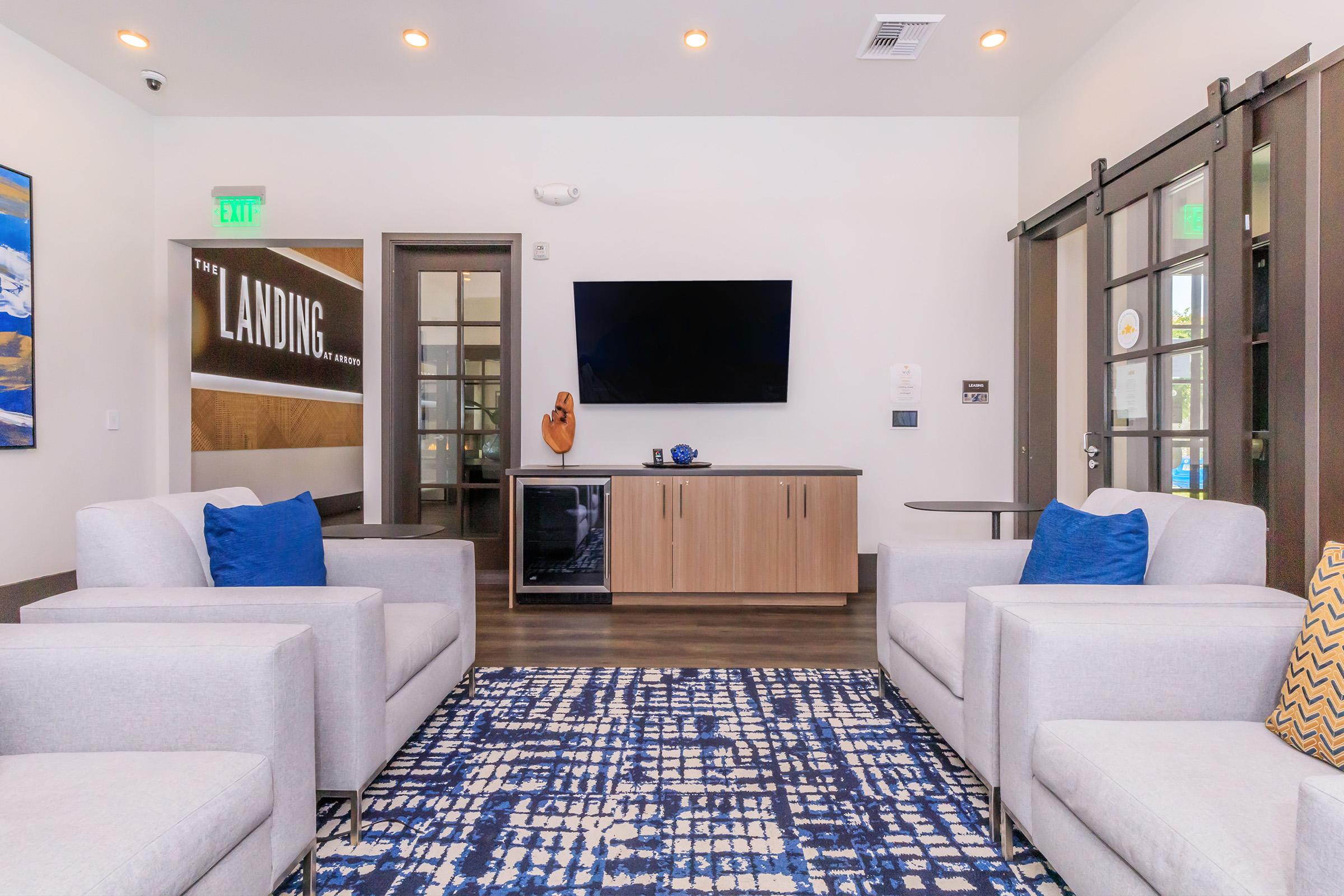A modern lounge area featuring two light gray armchairs with blue pillows, a wooden entertainment unit with a flat-screen TV, and a small table. The space is decorated with blue and white artwork on the walls, a decorative bowl on the unit, and a textured rug. Natural light illuminates the room from large windows.