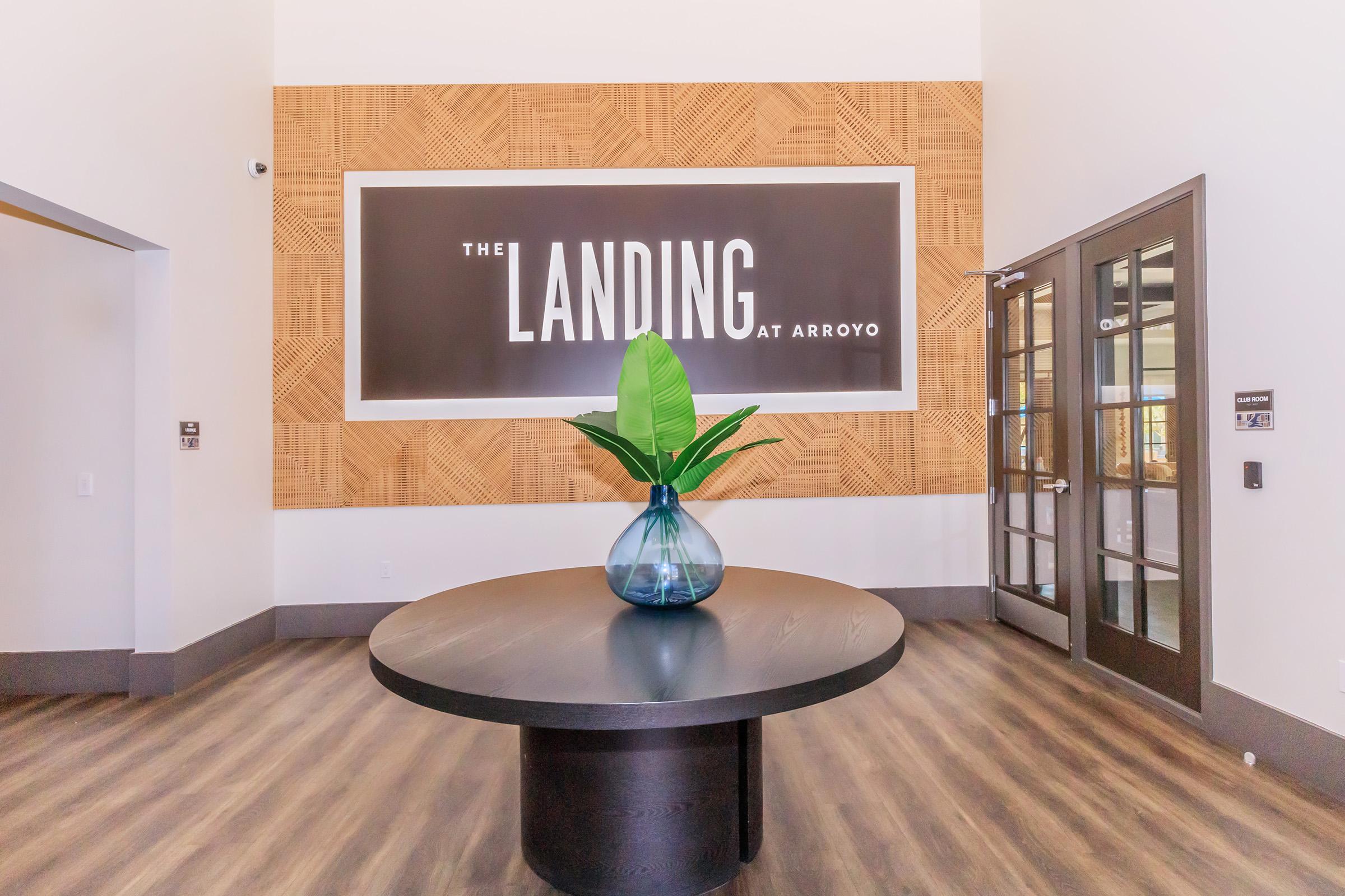 A modern lobby area featuring a large circular black table with a decorative blue vase and a green plant. Behind the table is a wall with the text "The Landing at Arroyo" displayed in bold white letters on a wood-patterned backdrop. The space has a contemporary design with wood flooring.