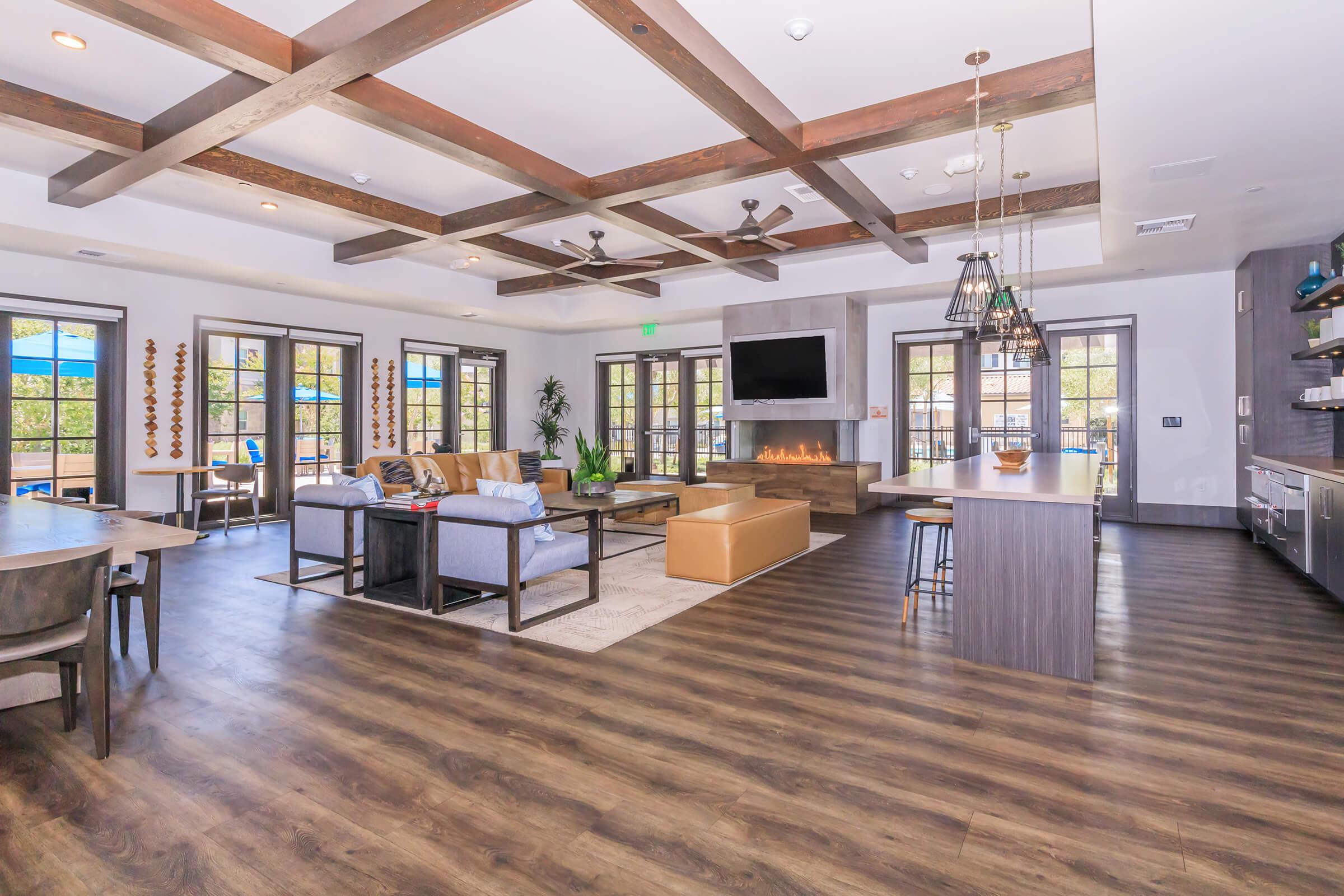A spacious, bright living area featuring wooden beams on the ceiling, large windows with natural light, a cozy sectional sofa, and a coffee table. There's a stylish kitchen area with modern appliances and an island, and a wall-mounted TV above a fireplace, adding a warm atmosphere.
