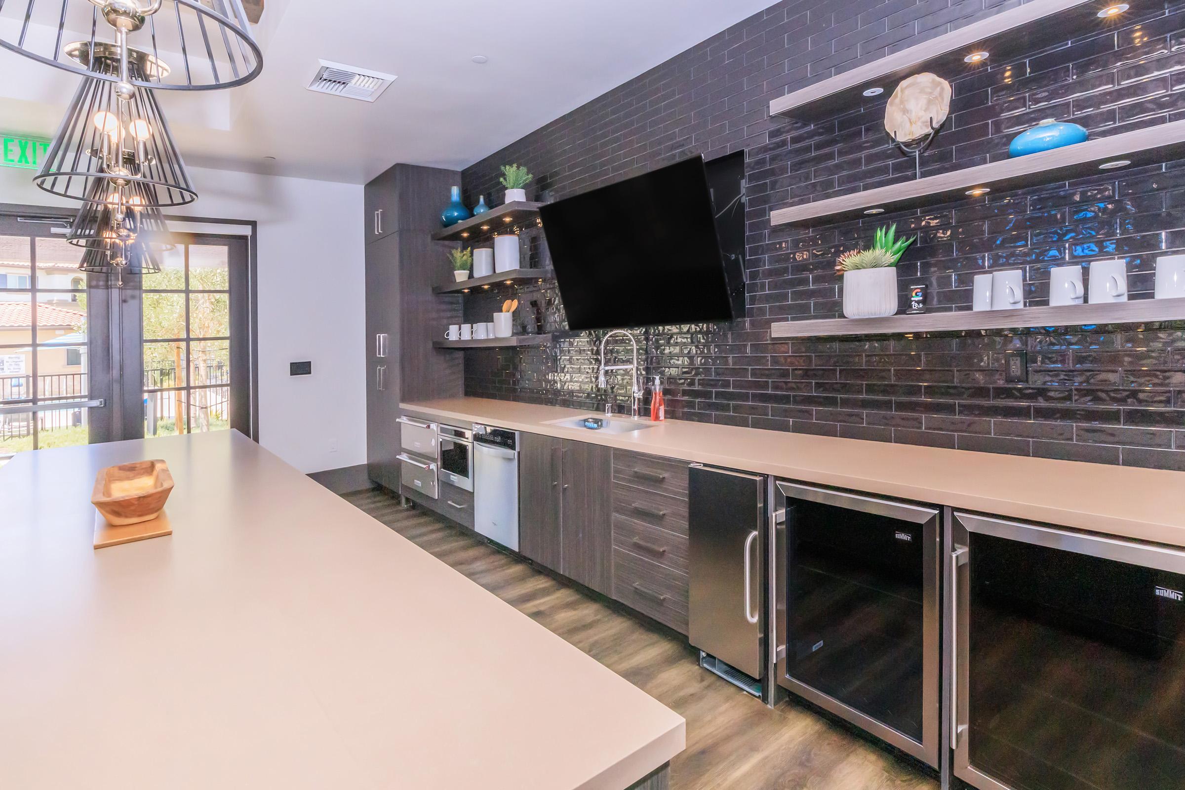 A modern kitchen showcasing sleek cabinetry and appliances. The space features a large countertop, a sink, and an under-counter refrigerator. Wall-mounted shelves display decorative items and plants. A large flat-screen TV is mounted on the wall, with a view of an outdoor area through glass doors.