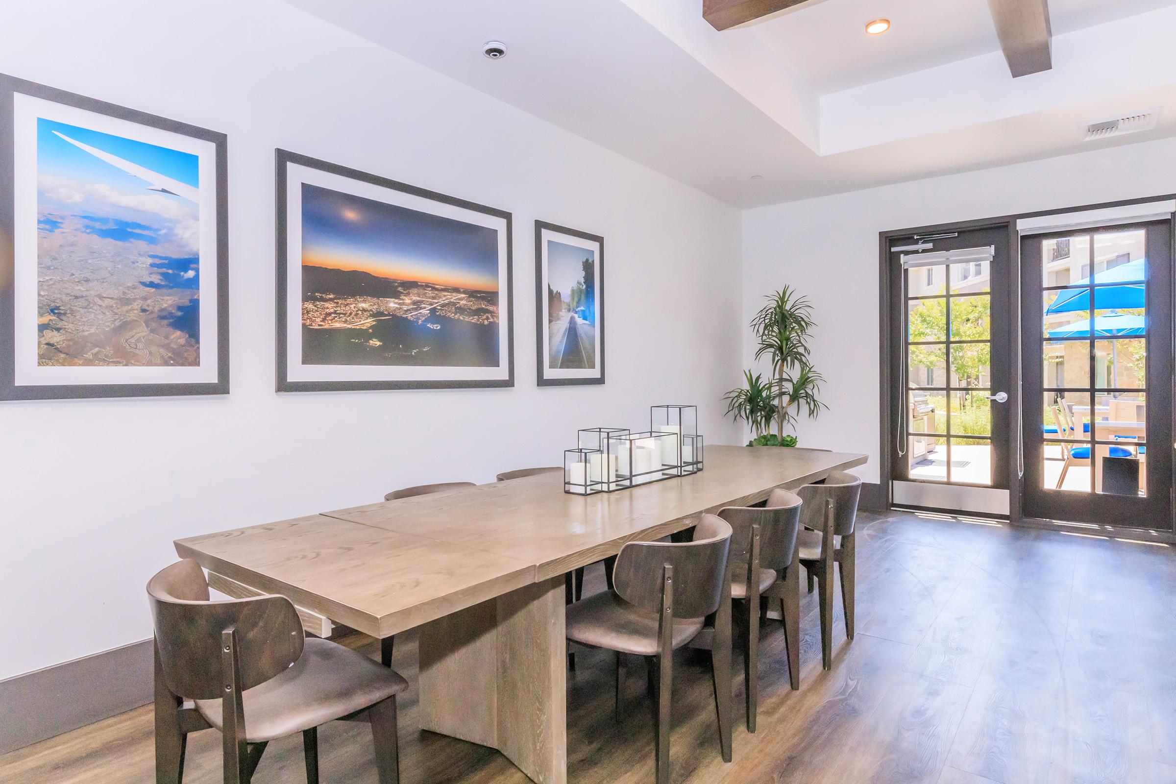 A modern dining area featuring a long wooden table surrounded by dark wooden chairs. The walls are adorned with three framed photographs showcasing landscapes and cityscapes. A large glass door leads outside, revealing a patio area with blue umbrellas and greenery. The space is bright and inviting.