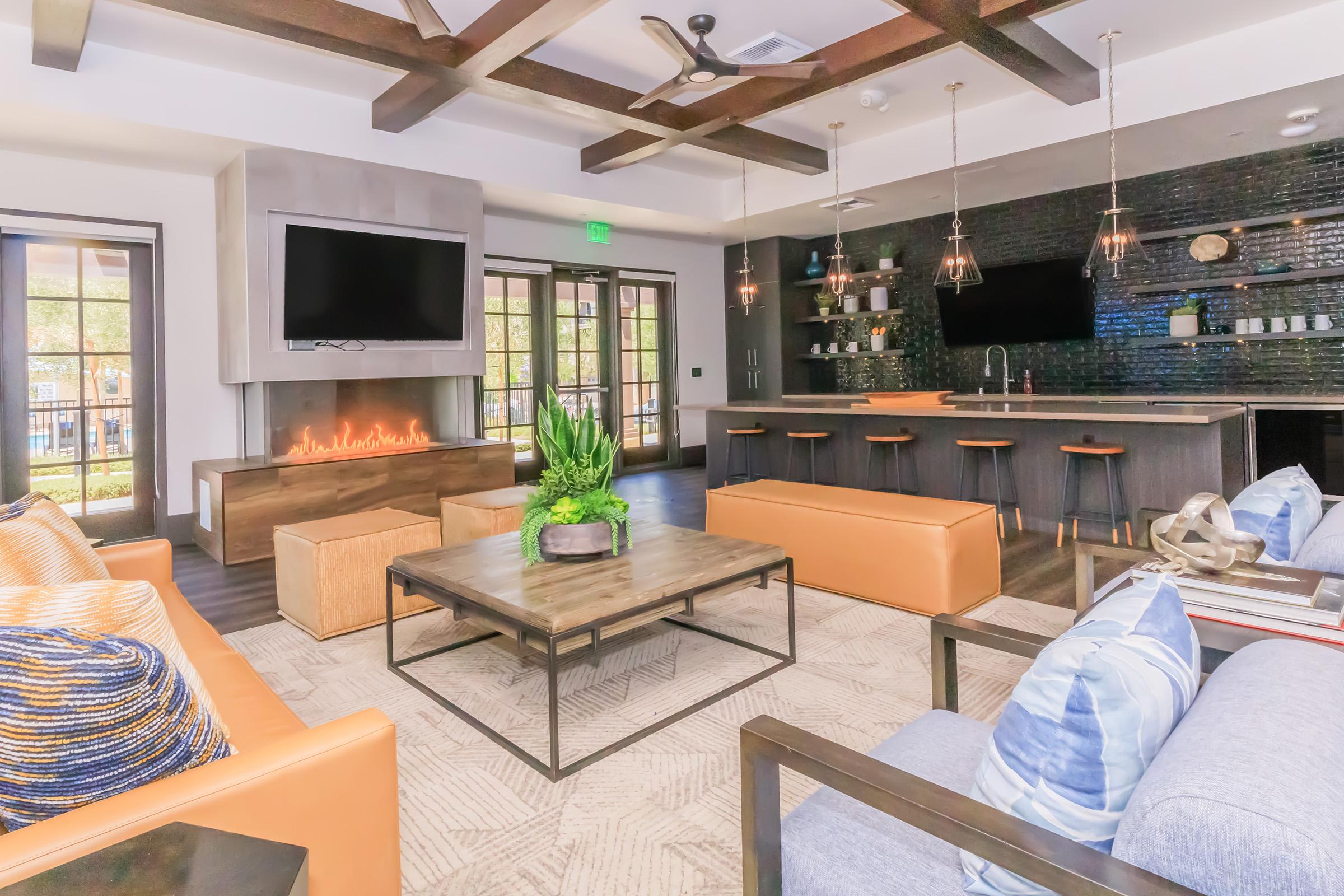 A modern living room featuring a cozy seating area with orange and blue sofas, a wooden coffee table, and a decorative plant. A flat-screen TV is mounted on the wall, and a stylish kitchen bar with pendant lighting is in the background, creating an inviting and contemporary atmosphere.