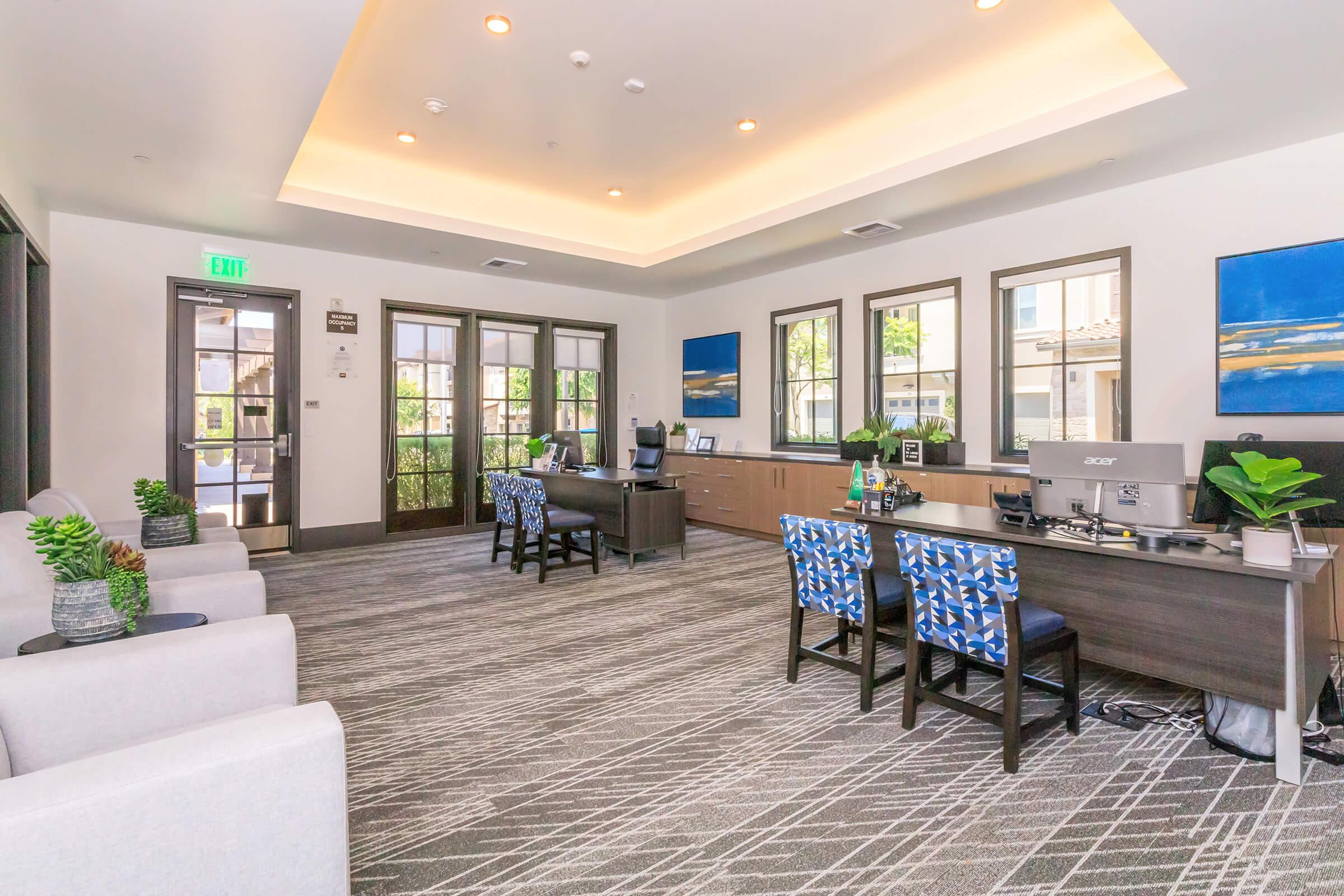 Interior view of a modern office space featuring a reception area with gray furniture, blue accent chairs, and desk equipped with computer and decorative plants. Large windows allow natural light in, creating a bright atmosphere. Artwork adorns the walls, adding color to the space.