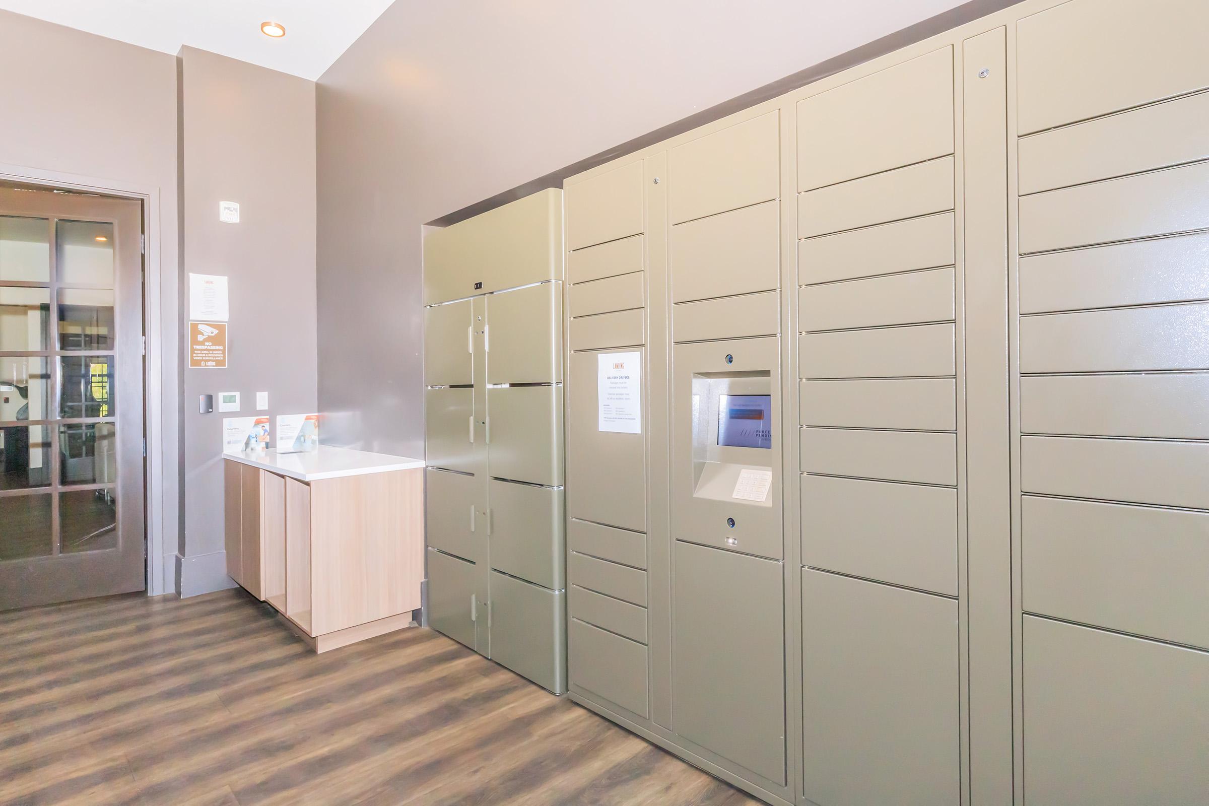 Interior view of a modern parcel locker area featuring several green lockers arranged in a wall. A countertop with brochures is visible to the left. The floor is made of wood-like material, and the walls are painted in neutral tones, creating a sleek and contemporary atmosphere.