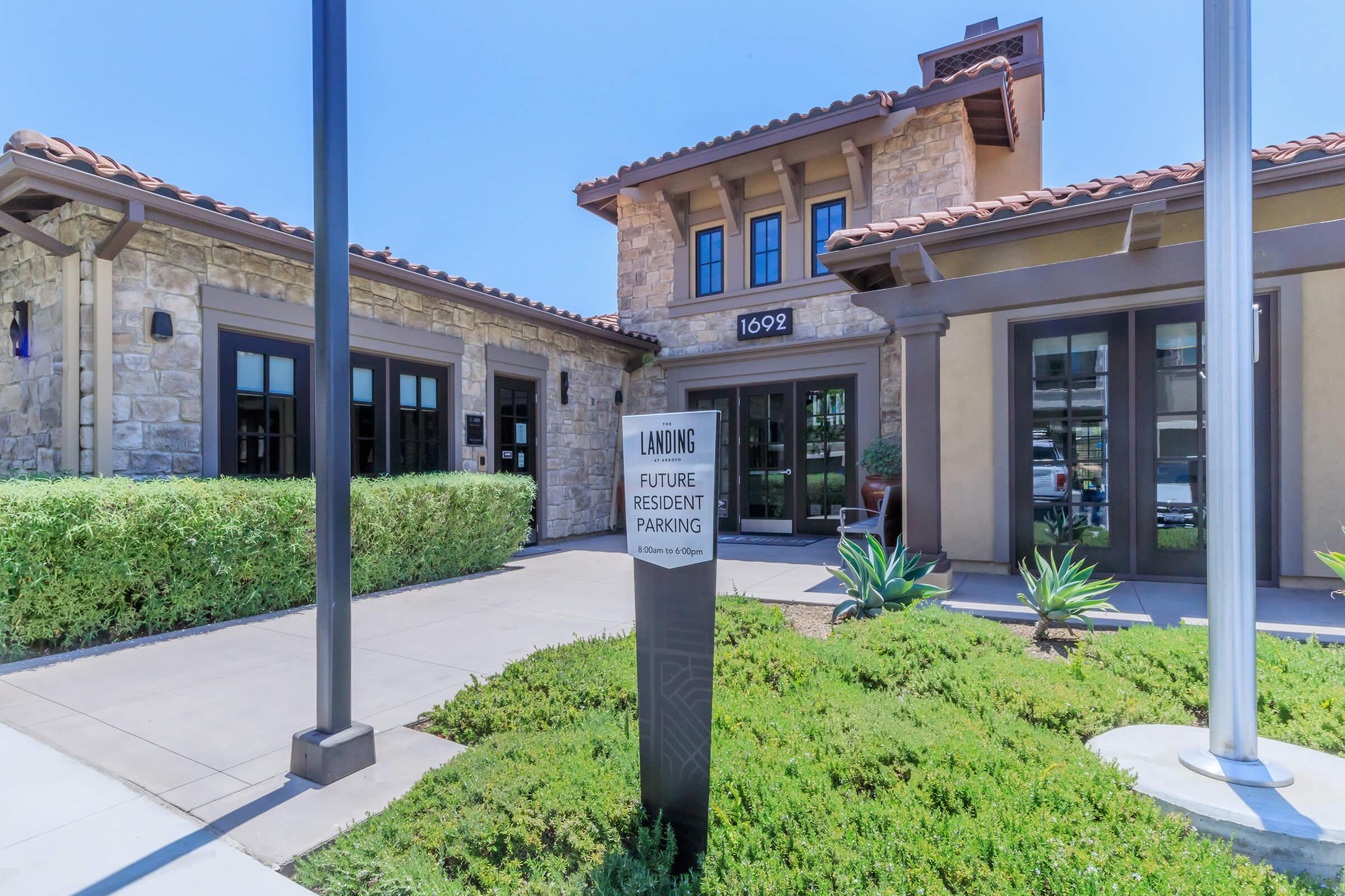 Building exterior of a residential complex featuring a stone facade and tiled roof. A sign reading "Landing Future Resident Parking" is prominently displayed in front. Lush greenery and ornamental plants surround the entrance, with clear blue skies above.