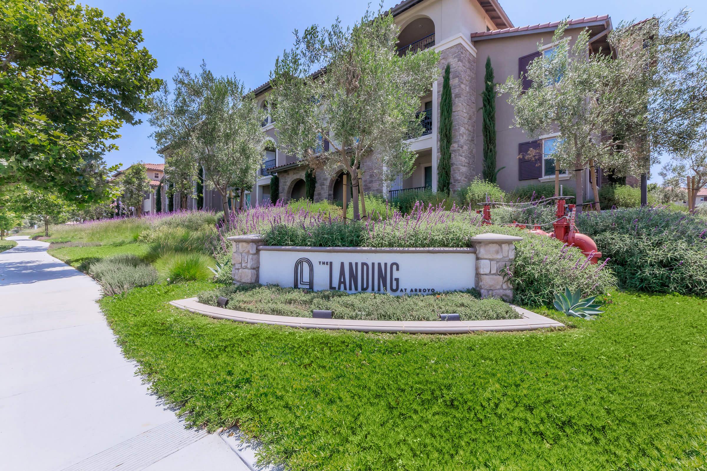 A landscaped entrance to an apartment complex named "The Landing." The sign is prominently displayed in front of well-maintained greenery and flowering plants, with residential buildings visible in the background under a clear blue sky.
