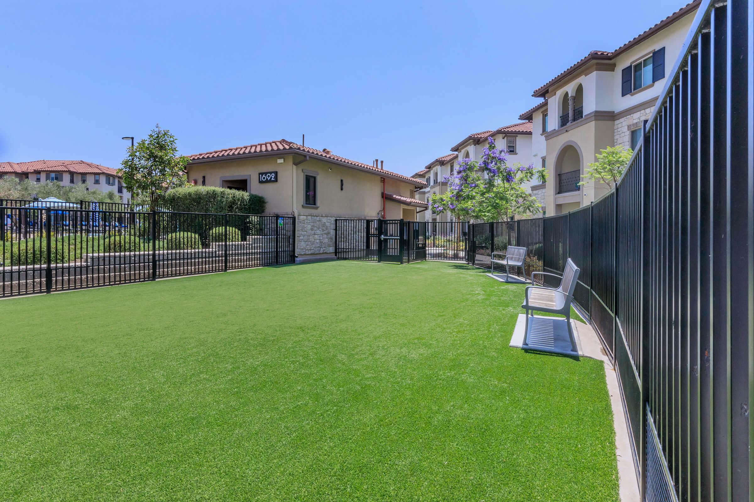 A well-maintained, fenced dog park with synthetic grass. Several benches are positioned along the side, and the surrounding area features landscaped shrubs and trees. In the background, there are residential buildings with a Mediterranean architectural style. Clear blue sky overhead.