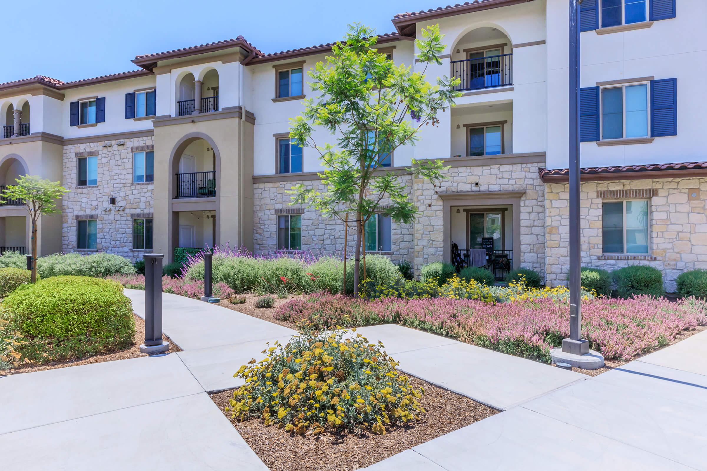 A sunny residential complex featuring a two-story building with stone accents, arched entrances, and balconies. The landscaped area includes a small tree, colorful flowering plants, and neatly trimmed shrubs, with a paved path leading to the entrance.