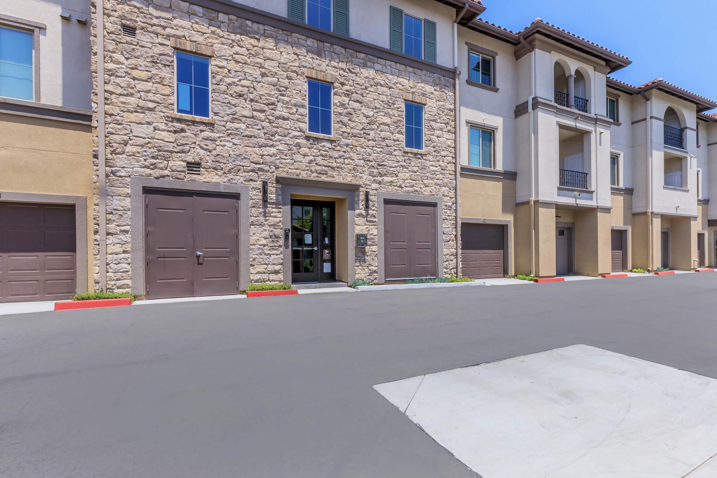 Modern multi-unit residential building featuring a stone façade and balconies. The entrance is framed by large windows and is flanked by garage doors. The surrounding area is paved, with landscaped borders enhancing the exterior. The clear blue sky suggests a bright, sunny day.