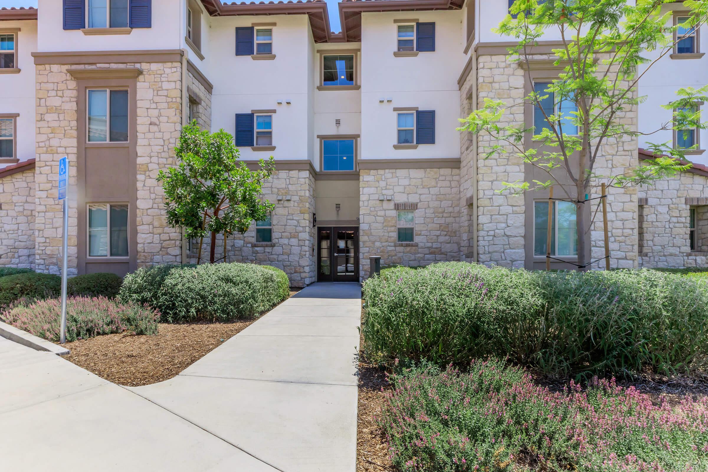 A modern multi-story building with a stone facade, featuring large windows and a welcoming entrance. Lush greenery and flowering plants line the pathway leading up to the entrance, creating a pleasant outdoor environment. The area is well-kept and inviting, with a clear sidewalk and landscaped shrubs.