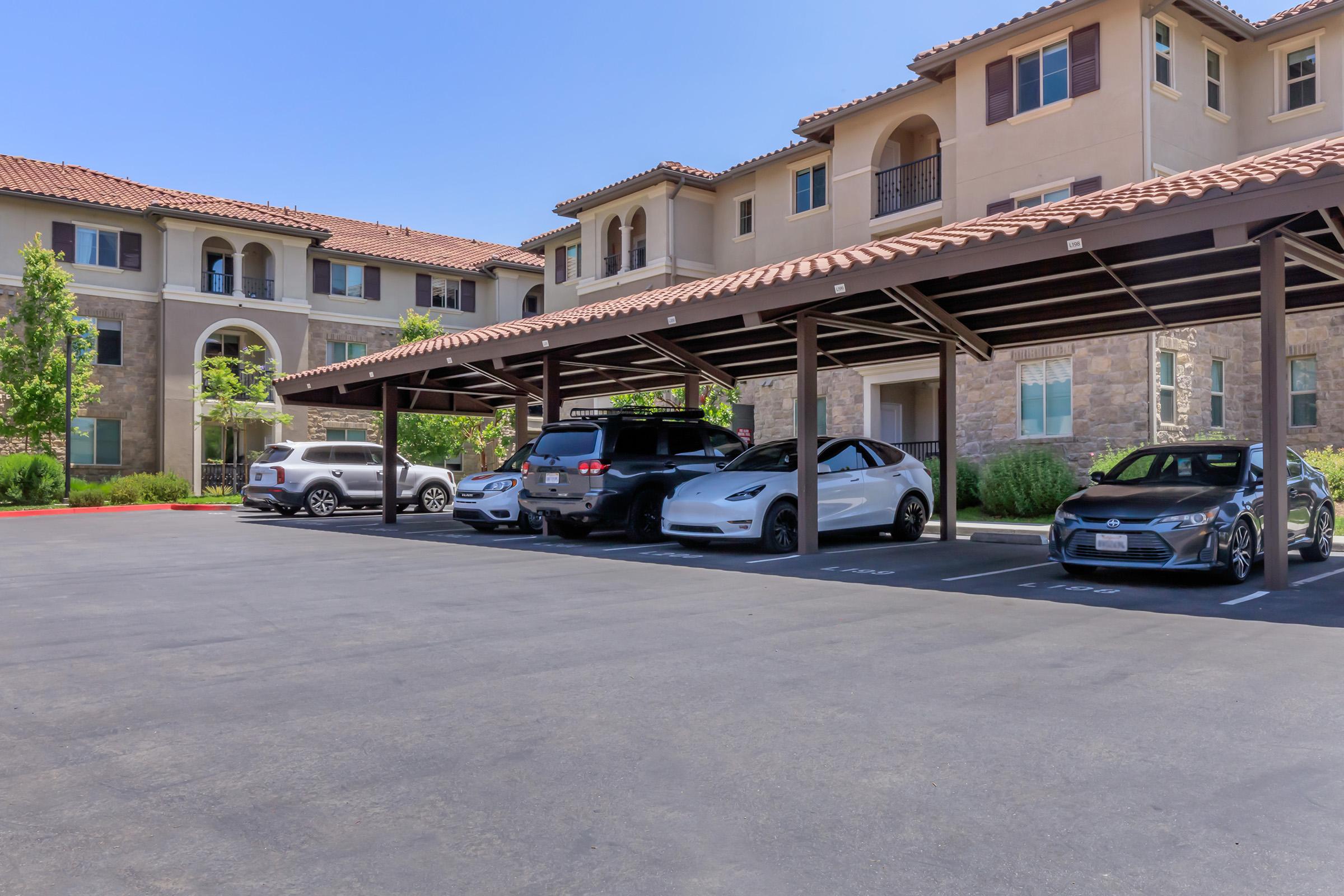 Row of cars parked under a shaded carport in front of a multi-story apartment building. The building features a stone facade and tile roof, with green trees and blue sky in the background.