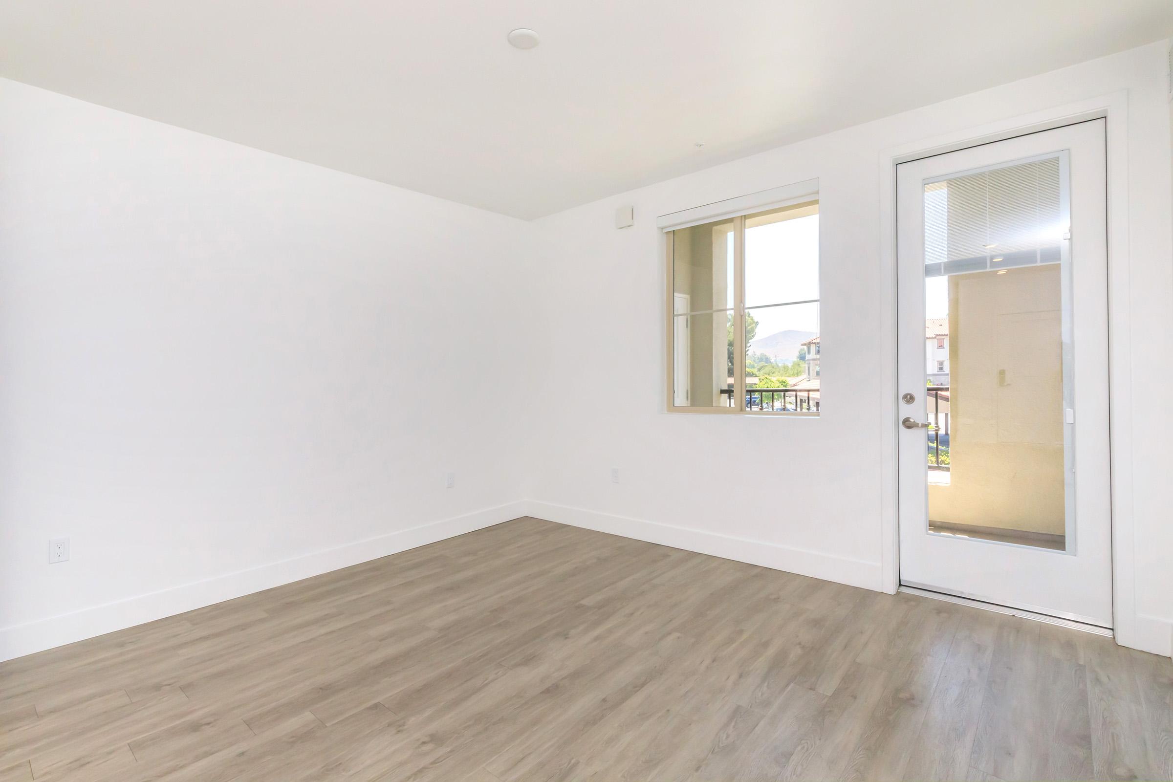 A bright, empty room with white walls and light wooden flooring. A large window provides natural light, and a sliding glass door leads to a balcony, showcasing a view of greenery outside. The room has minimal furnishings, creating a spacious and airy atmosphere.