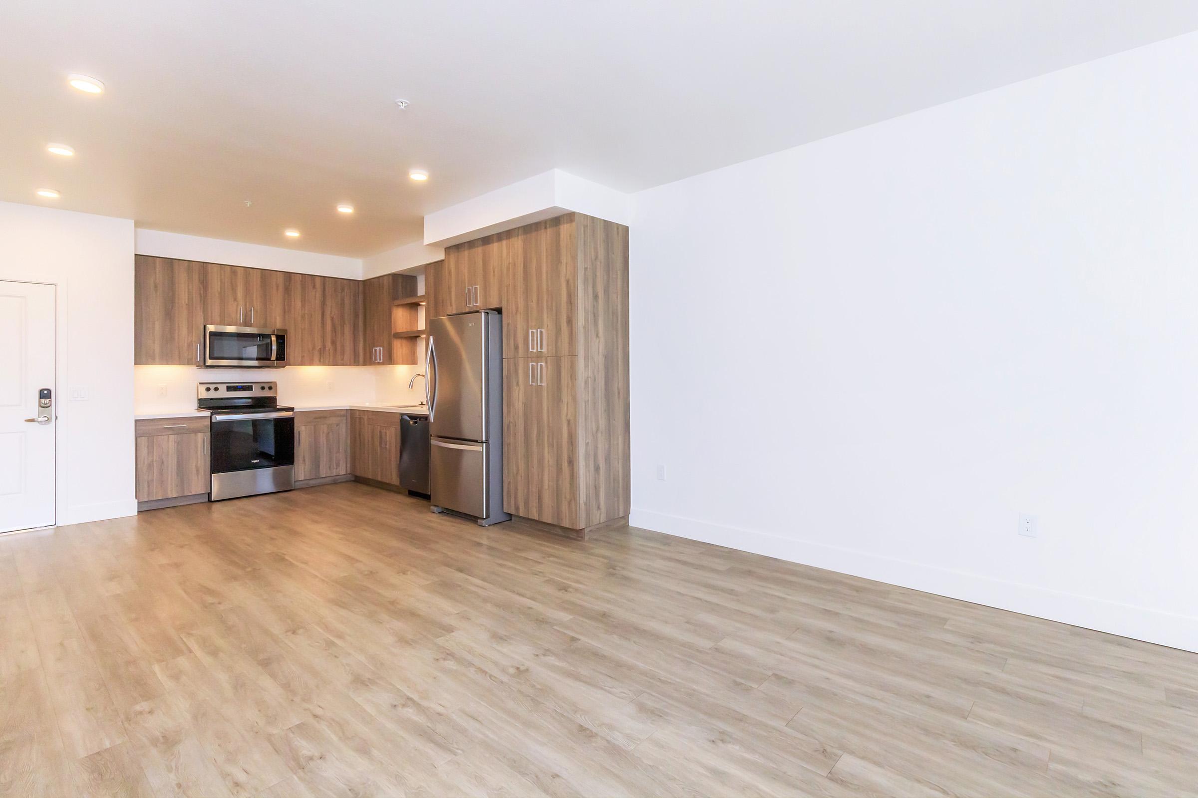 Modern kitchen and living area featuring wooden cabinetry, stainless steel appliances, and a spacious layout with light wood flooring. Bright, neutral walls enhance the open space, allowing for versatile furniture arrangements. Ideal for contemporary living.