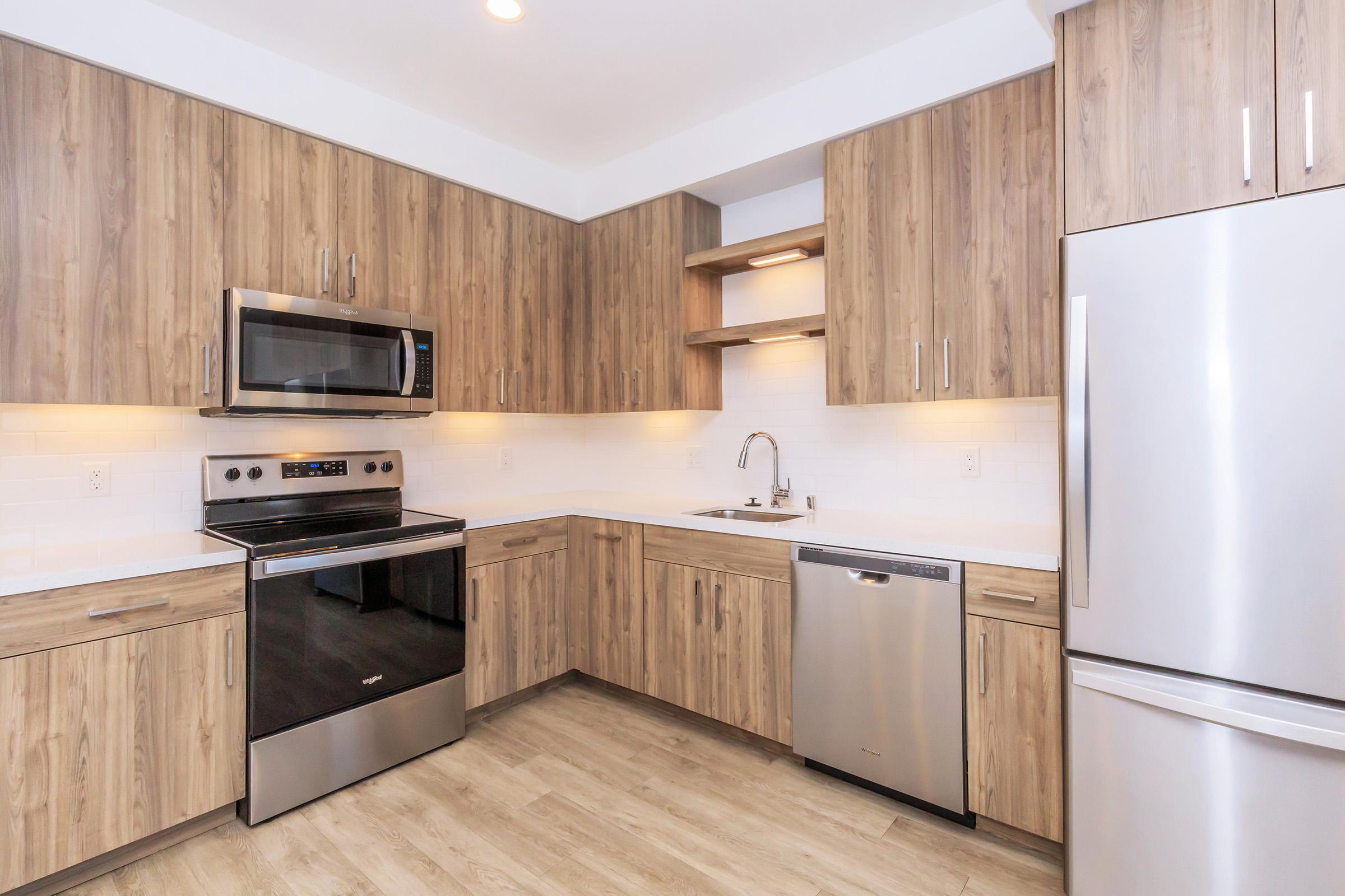 Contemporary kitchen with wooden cabinetry and stainless steel appliances. Features an oven, microwave, dishwasher, and a sink. The countertop is light-colored, and there are under-cabinet lights illuminating the workspace. The flooring is a light wood finish, creating a modern, cohesive look.