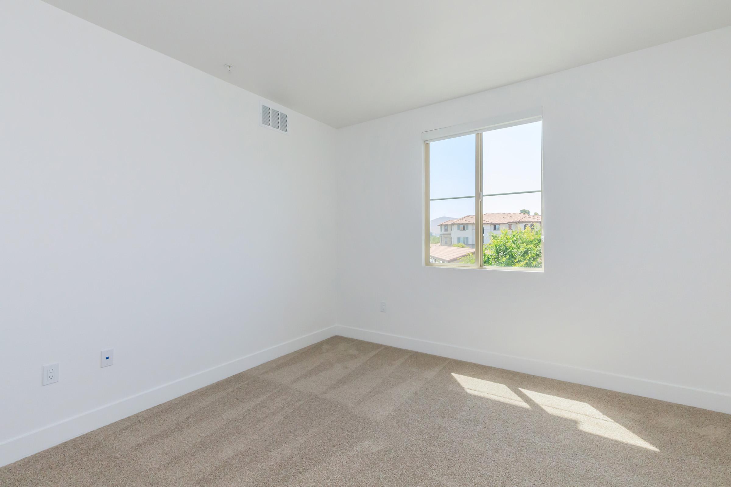 A bright, empty room with white walls and a large window allowing natural light to enter. The floor is covered in light-colored carpet, and there are no furnishings or decorations. Outside the window, you can see the greenery of trees and a glimpse of buildings.