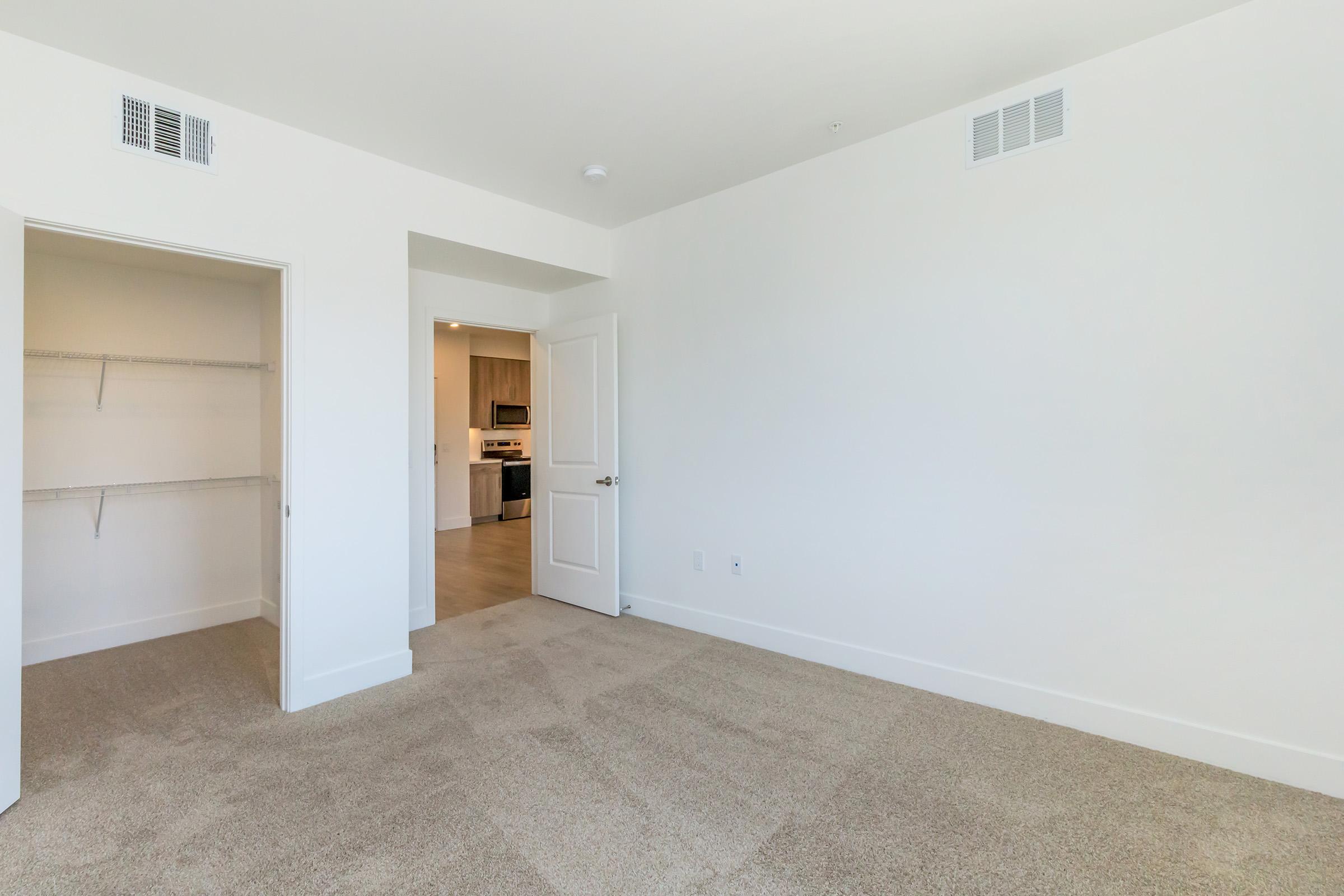 A spacious, empty bedroom with light-colored walls and carpet. One side features a closet with shelves, while a doorway leads to a kitchen area in the background. The room is well-lit with natural light, providing a clean and modern feel suitable for personal decoration.