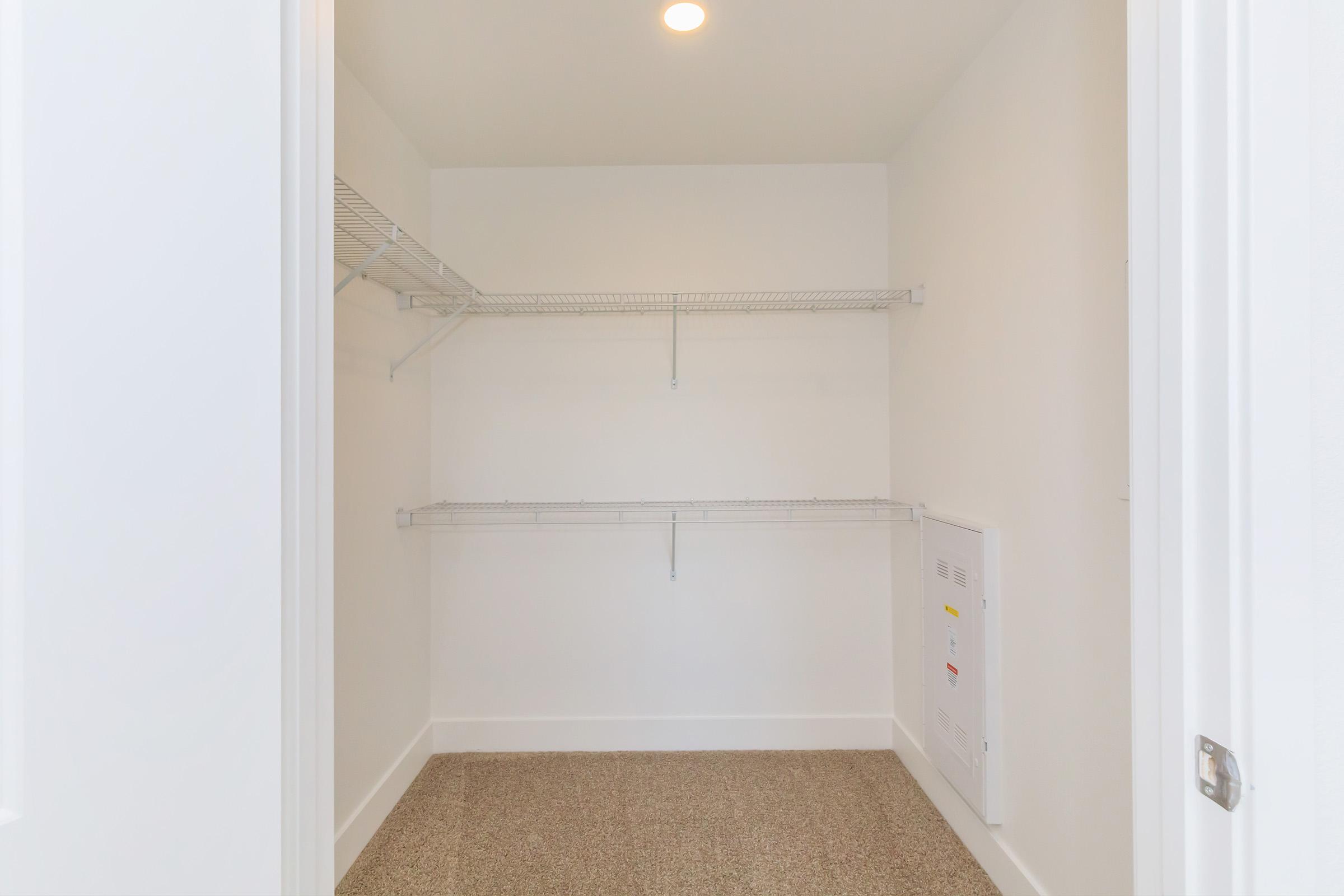 Empty walk-in closet featuring white walls and two wire shelves on opposite sides. The floor is a light-colored textured surface, and there is a small utility panel on the right wall. A ceiling light provides illumination, creating a bright and spacious appearance.