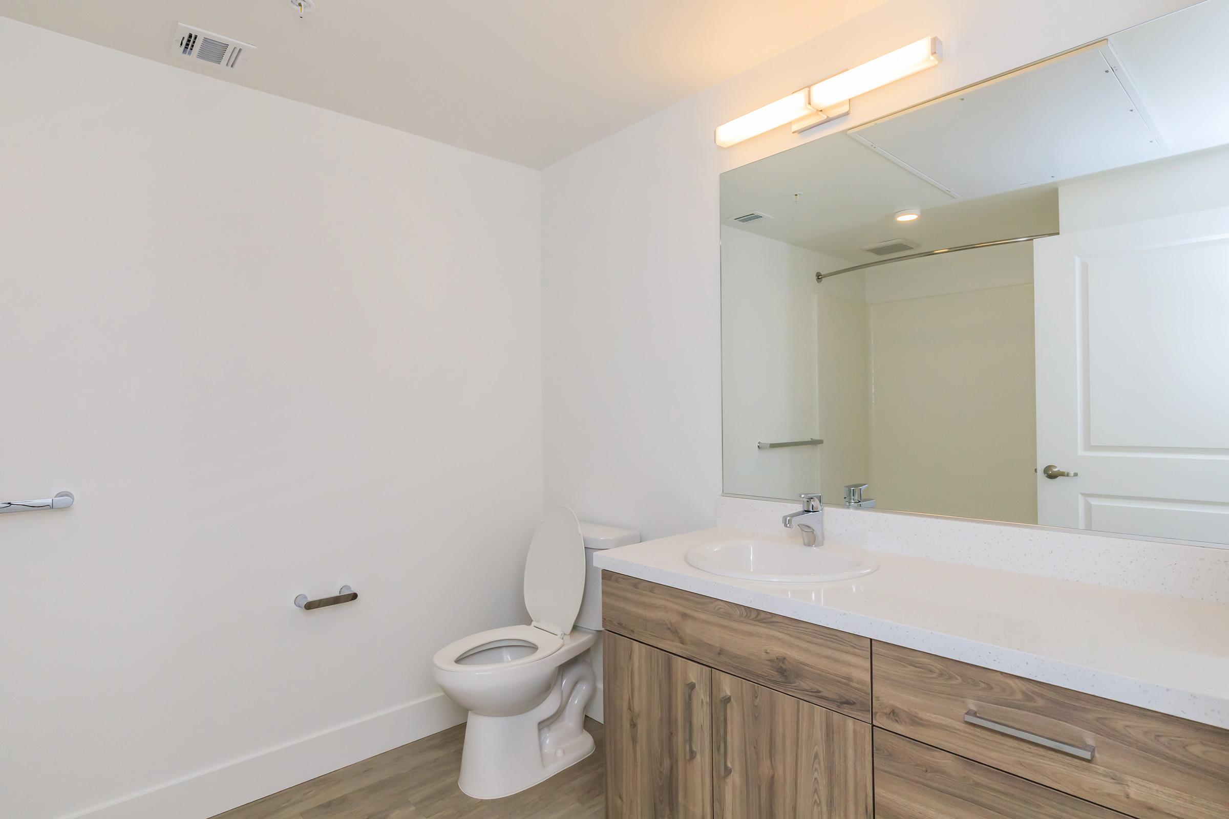 A clean, modern bathroom featuring a white toilet, a wooden vanity with a sink, and a large mirror. The walls are painted white, and the flooring has a light, wooden finish. A towel bar is mounted on the wall, and there is a partially mirrored cabinet above the sink.