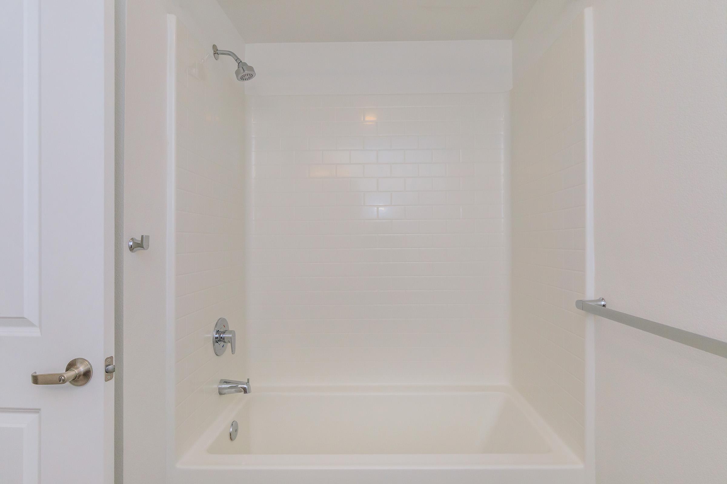 A clean, empty bathroom featuring a white tiled shower and bathtub. The walls are smooth and lightly illuminated, with a showerhead mounted on the wall. There is a towel bar on one side, and a white door is slightly visible in the frame. The overall atmosphere is bright and minimalist.