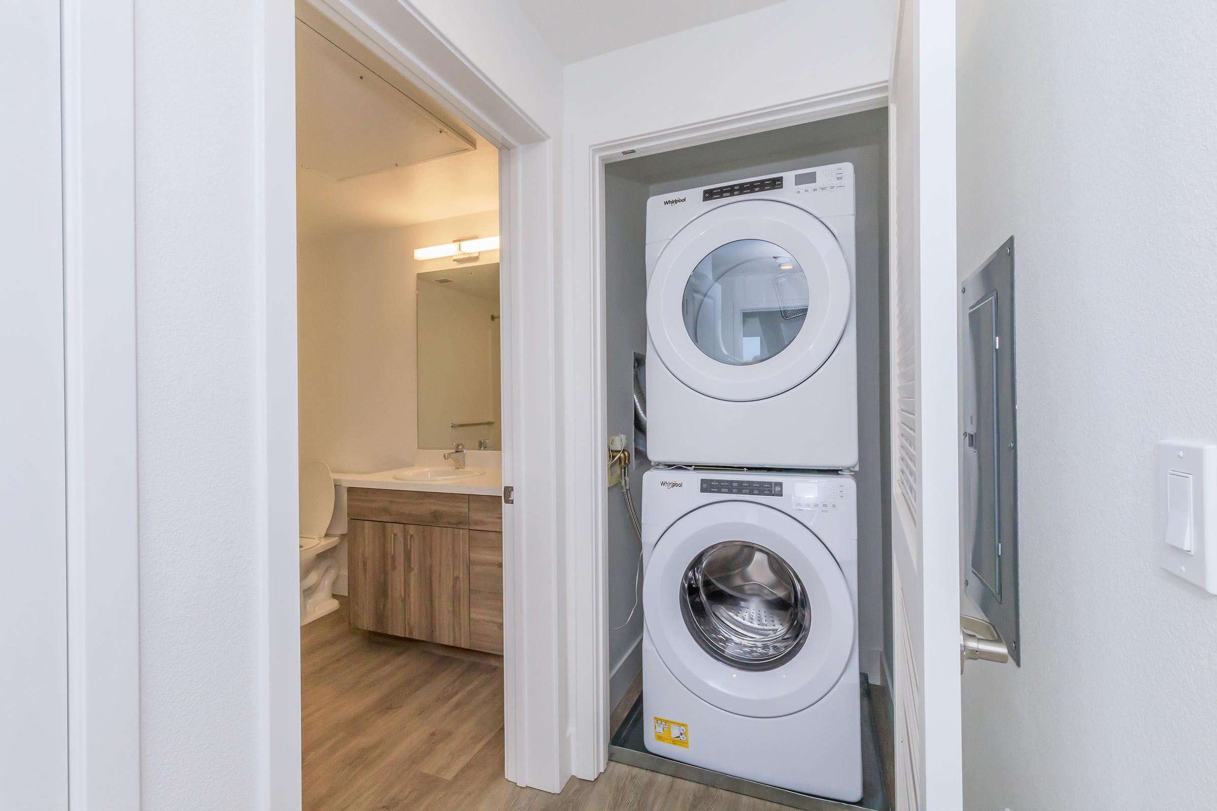 Stacked washer and dryer located in a narrow laundry area, adjacent to a bathroom with a sink and mirror. The space features light-colored walls, wooden cabinetry, and modern appliances, with a door leading to the bathroom.