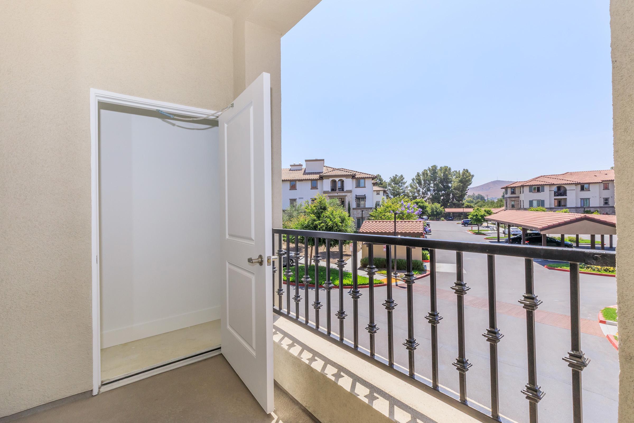 View from a balcony showing an open door, leading to a small storage area. The scene includes a sunny sky and a residential area with buildings and parked cars visible in the background. The railing is decorative, enhancing the balcony's aesthetic.