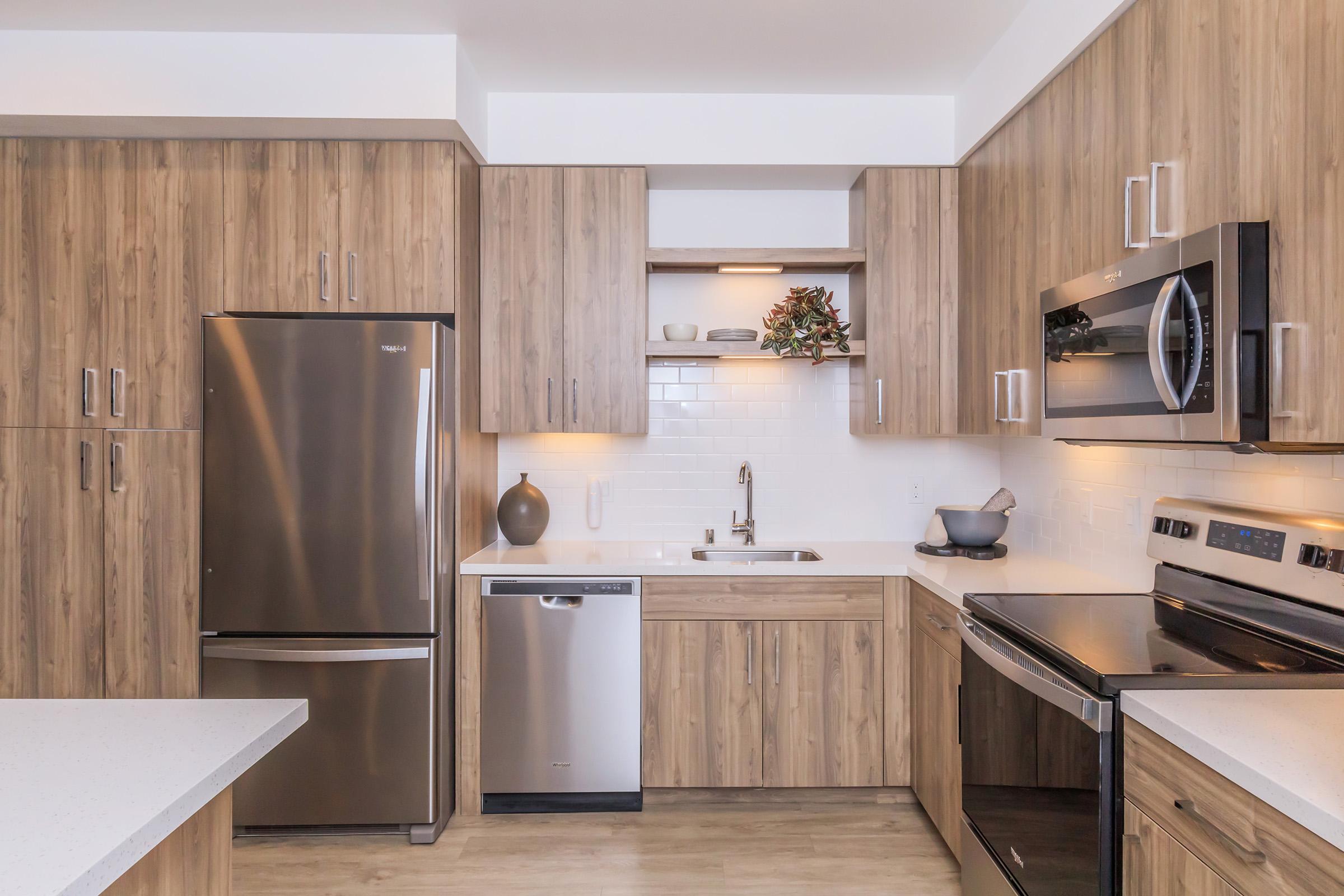 Modern kitchen featuring wooden cabinetry, stainless steel appliances including a refrigerator, dishwasher, and oven, as well as a microwave. The countertops are light-colored, and the space is well-lit, with decorative items on shelves above the sink. The flooring is a light wood laminate.