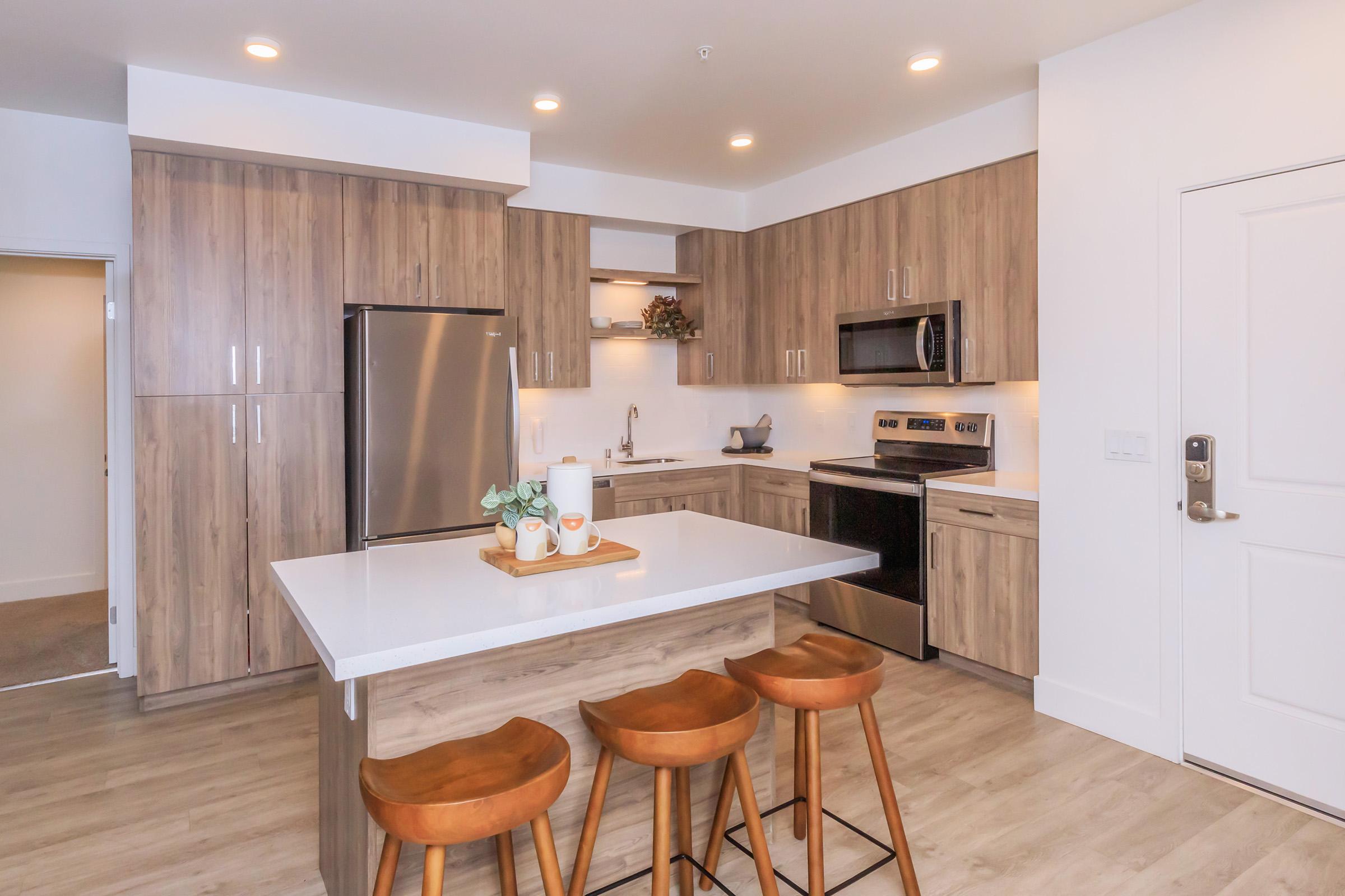 Modern kitchen featuring wooden cabinetry, stainless steel appliances, and a large island with seating for three. The space is well-lit with recessed lighting, and decorative items are placed on the countertop, adding a cozy touch to the contemporary design. A door leads to another room.