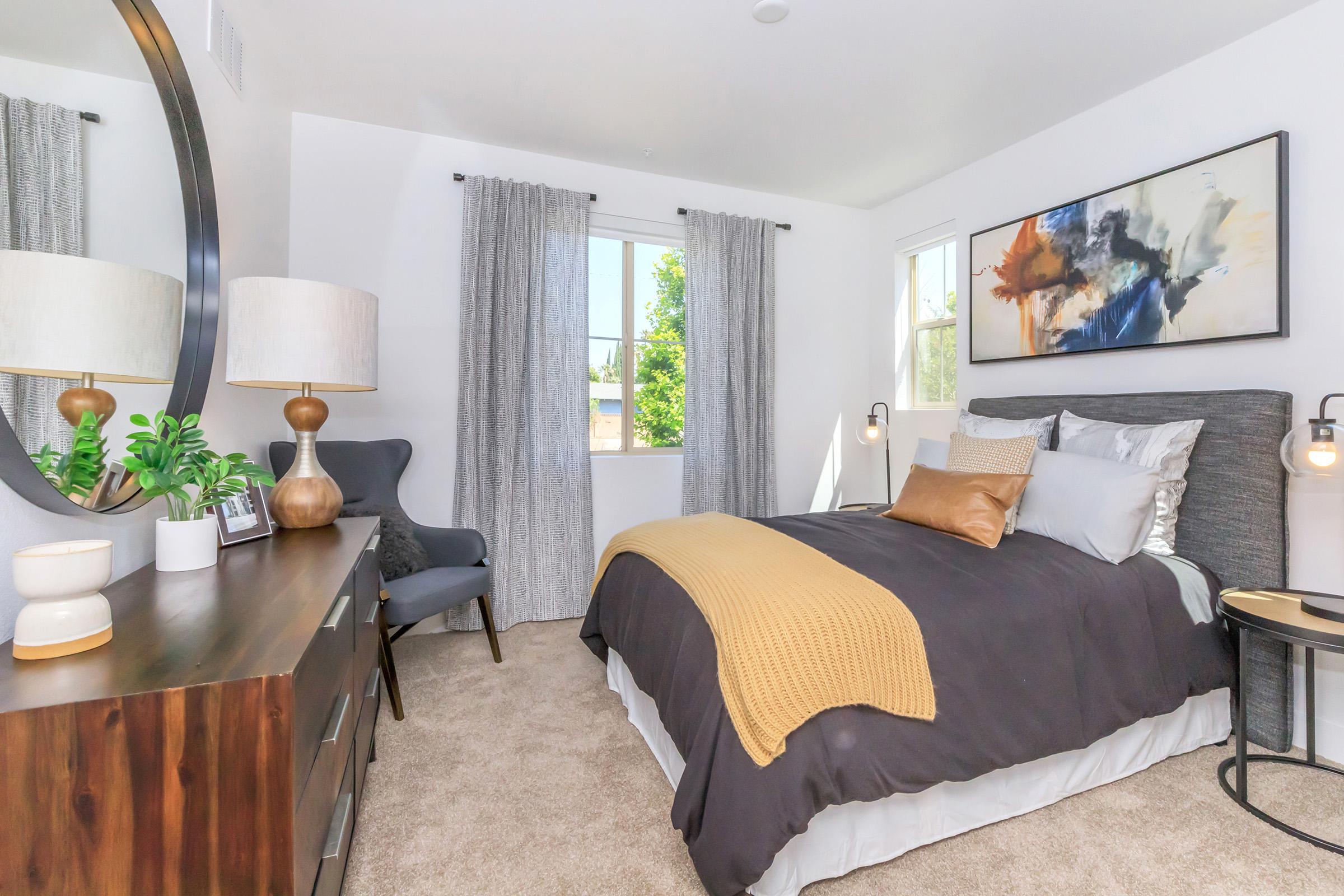 A cozy bedroom featuring a comfortable bed with dark gray and white bedding, a mustard-colored throw blanket, and decorative pillows. There’s a stylish gray armchair in the corner, a round mirror above a wooden dresser, and a small nightstand with a lamp. Natural light flows in through a window with light gray curtains.