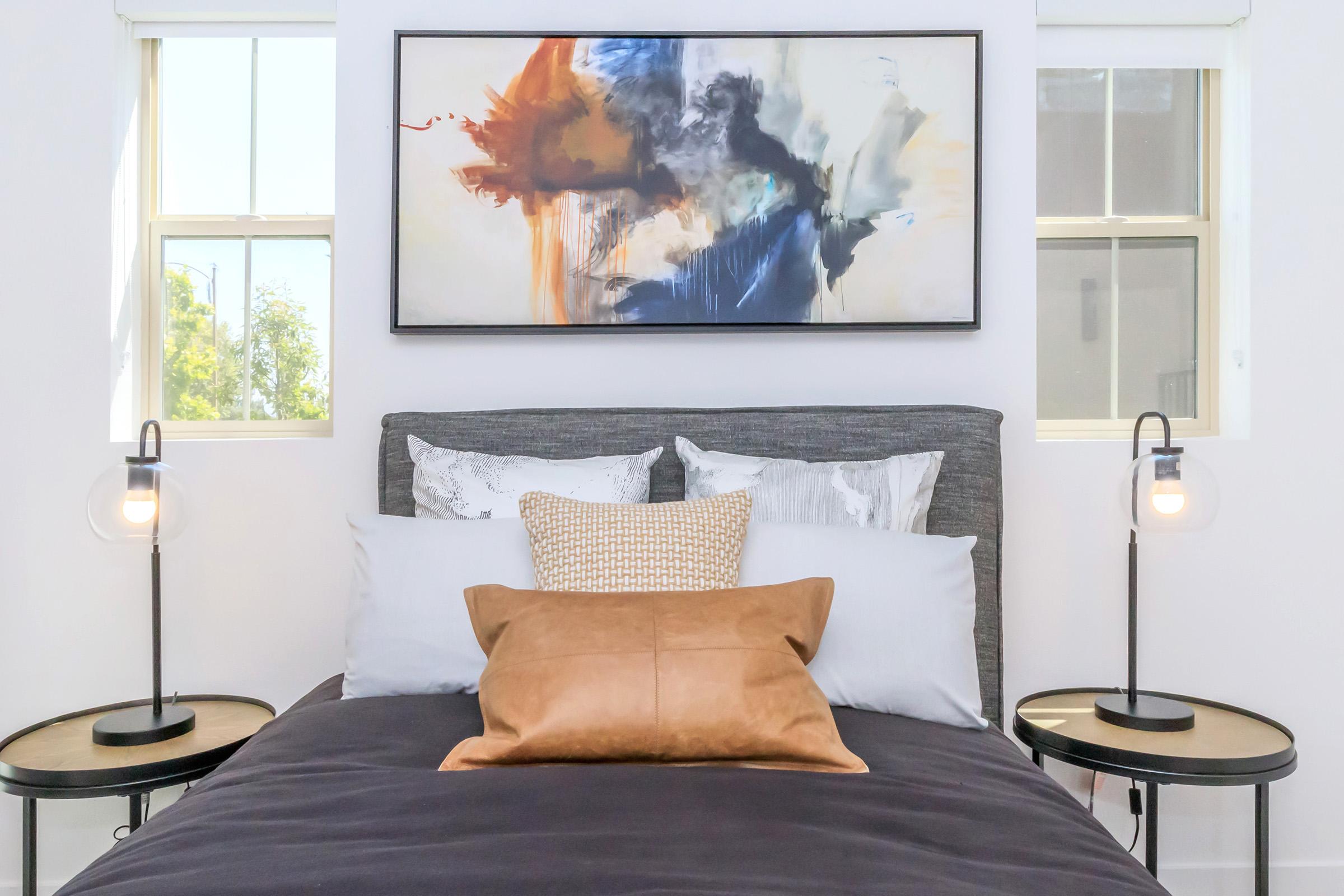A modern bedroom featuring a bed with multiple pillows, a brown cushion, and stylish lamps on small side tables. Above the bed hangs a large abstract painting with vibrant colors. Two windows provide natural light, enhancing the inviting atmosphere.