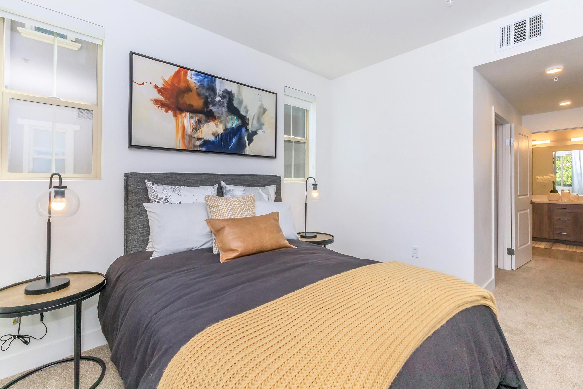 A modern bedroom featuring a cozy bed with dark bedding, decorative pillows, and a golden throw. Two stylish side tables with lamps are placed beside the bed. A colorful abstract painting hangs above the headboard, and a doorway leads to a well-lit bathroom area. Natural light fills the room from large windows.