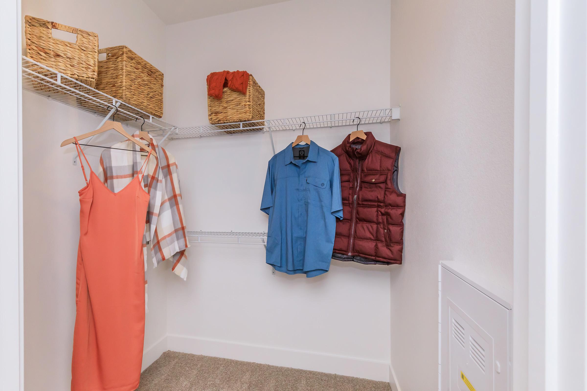 A walk-in closet featuring a hanging orange dress, a blue shirt, a burgundy vest, and a plaid scarf, along with two woven baskets placed on the top shelf, all set against white walls and a carpeted floor. The closet has a neat and organized appearance.