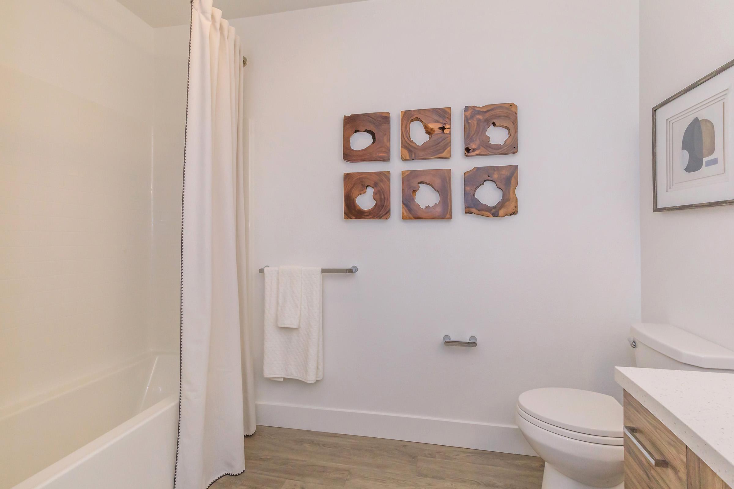A clean and modern bathroom featuring a bathtub on the left, a white shower curtain, and a towel rack with a neatly hung towel. On the wall, there are six wooden art pieces arranged in a grid, and a framed artwork is displayed next to the toilet and sink area. The floor is made of light-colored wood.