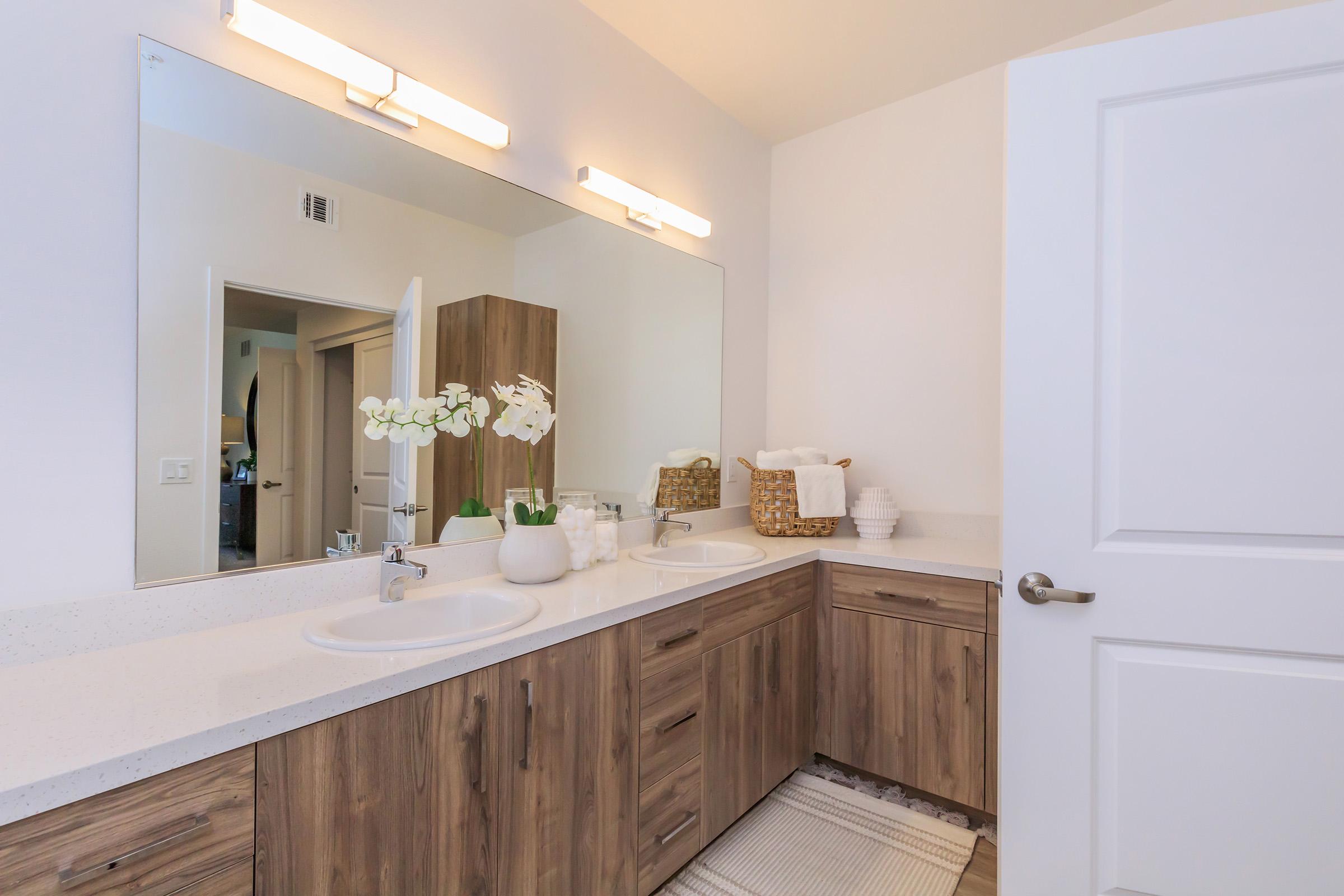 A modern bathroom featuring a double sink vanity with wooden cabinets, a large mirror above, stylish lighting, and decorative elements like orchids and towels. The clean, minimalist design emphasizes neutral colors and a spacious layout. A closed door leads to another area of the home.