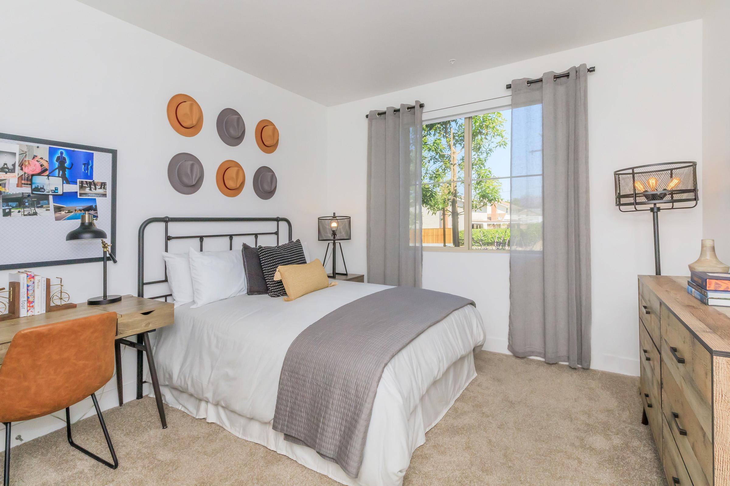 A cozy bedroom featuring a queen-sized bed with a gray and white bedding set. The walls are adorned with hats, and there are two curtains framing a window that lets in natural light. A stylish desk with a chair and decorative items are nearby, along with a dresser, creating a warm and inviting atmosphere.