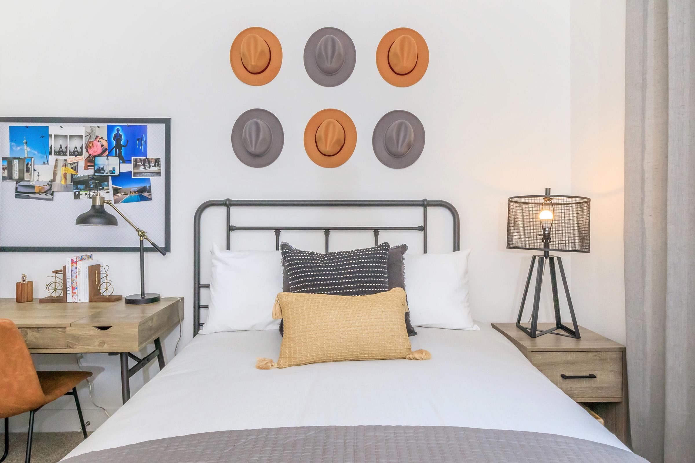 A cozy bedroom featuring a bed with white linens and decorative pillows, a stylish desk with a lamp, and a gallery wall displaying six hats. There is a bedside table with a lamp and a framed board containing photos and decor. Soft gray curtains frame a window, enhancing the room's inviting atmosphere.