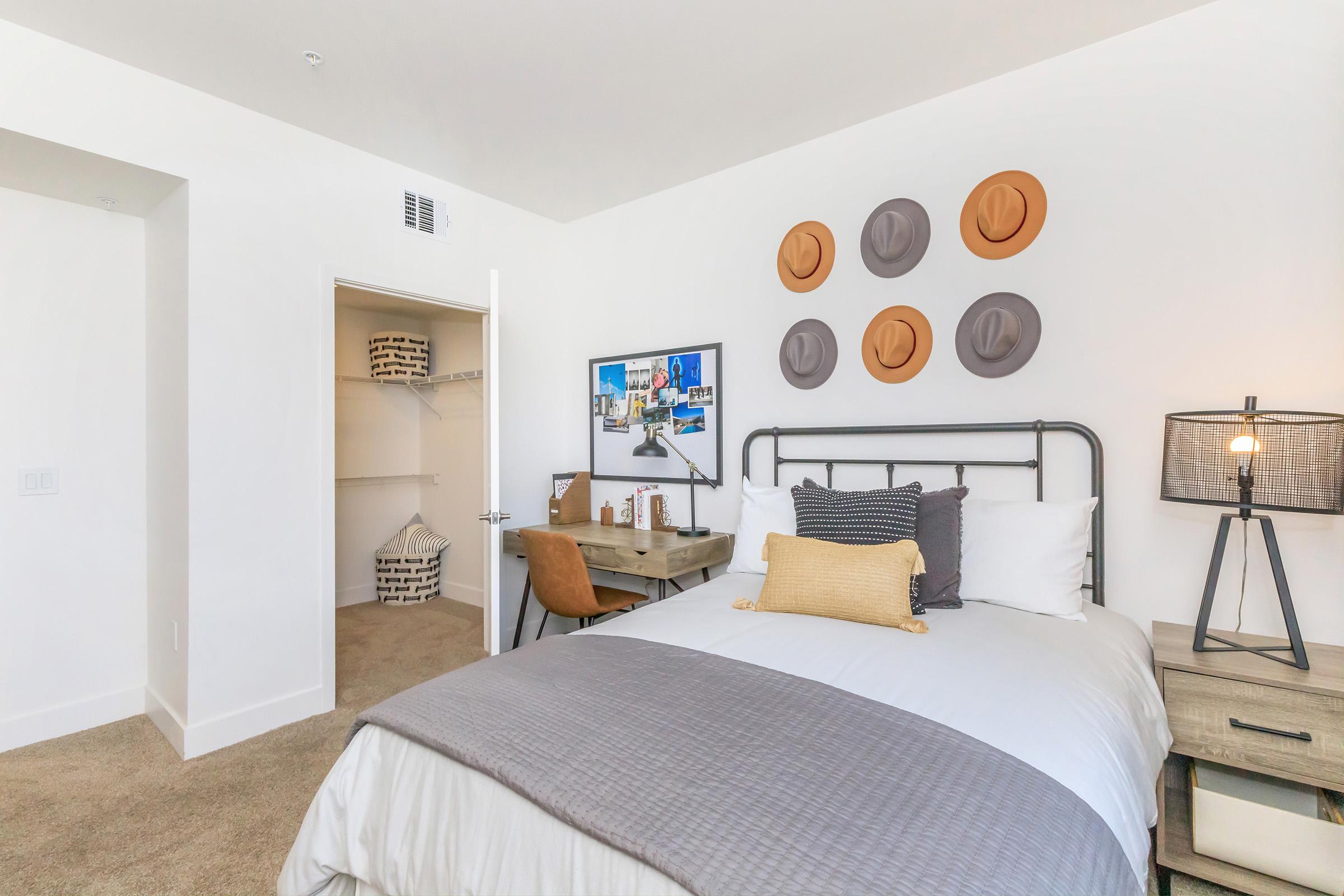 A well-decorated bedroom featuring a queen-sized bed with white linens, a gray throw blanket, and decorative pillows. A wall displays a mix of hats, while a framed picture is mounted nearby. There’s a desk with a lamp and a closet with storage baskets visible. The room has a modern and inviting aesthetic.