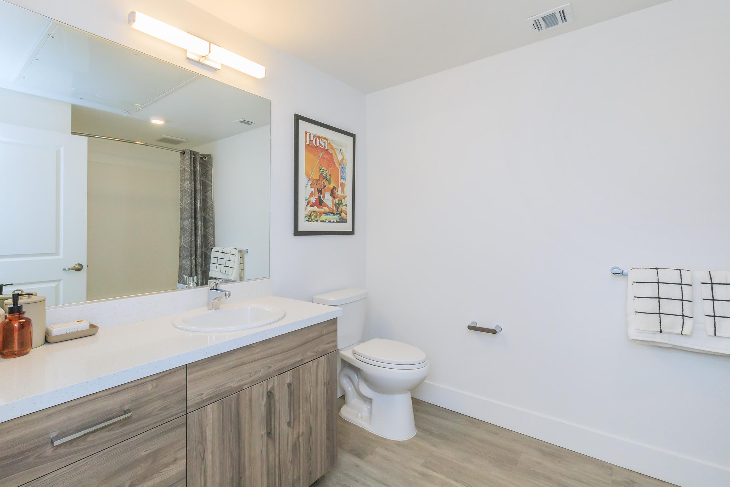 A modern bathroom featuring a white wall, a large mirror, and a wooden vanity with a sink. There is a towel rack with a white and black checkered towel, a toilet, and a decorative framed poster on the wall. Soft lighting illuminates the space, creating a clean and contemporary look.