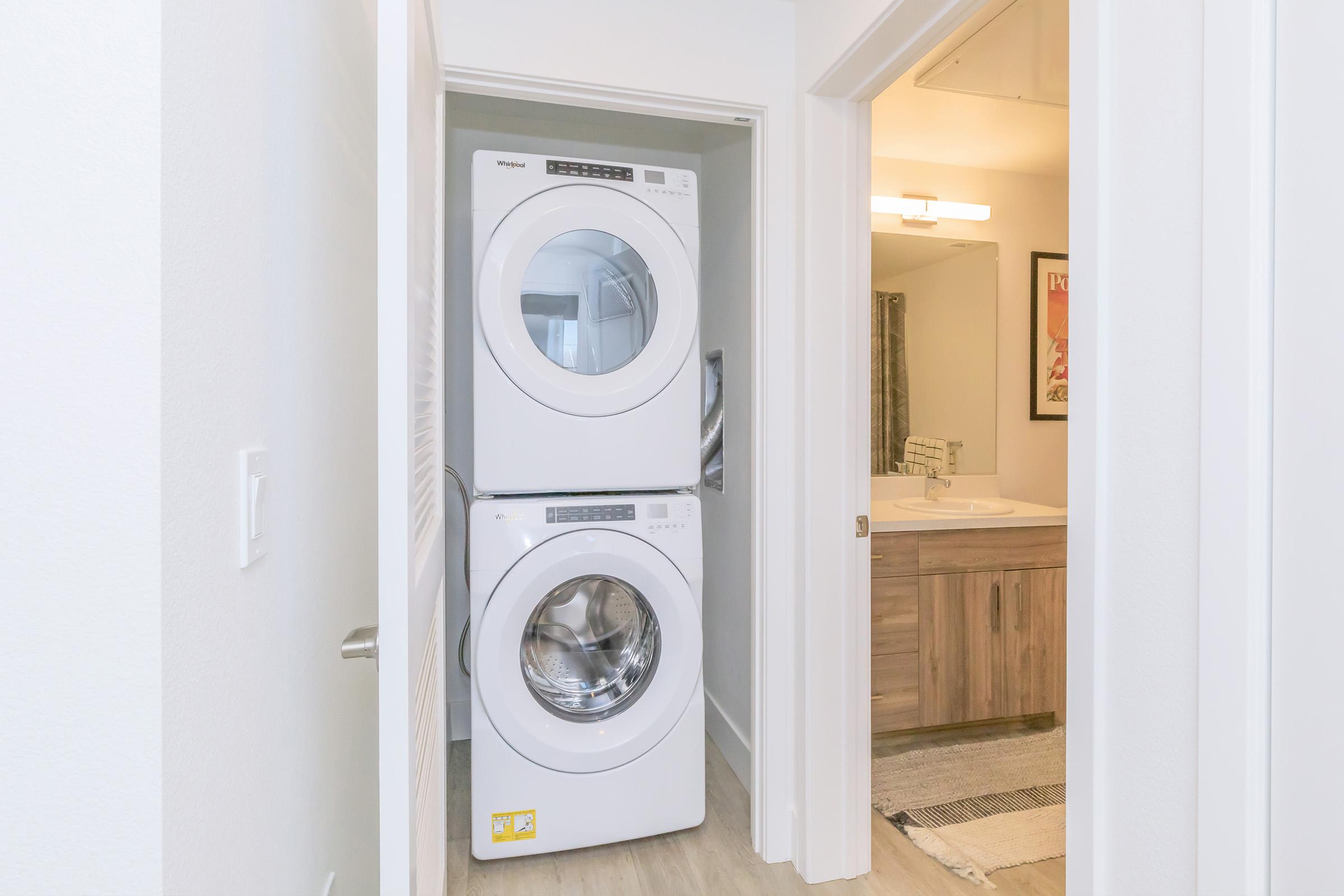 A laundry area featuring a stacked washer and dryer in a narrow space. The appliances are white and neatly arranged. In the background, a bathroom area is visible with light wood cabinetry and neutral-colored decor. The room has a bright, modern aesthetic.
