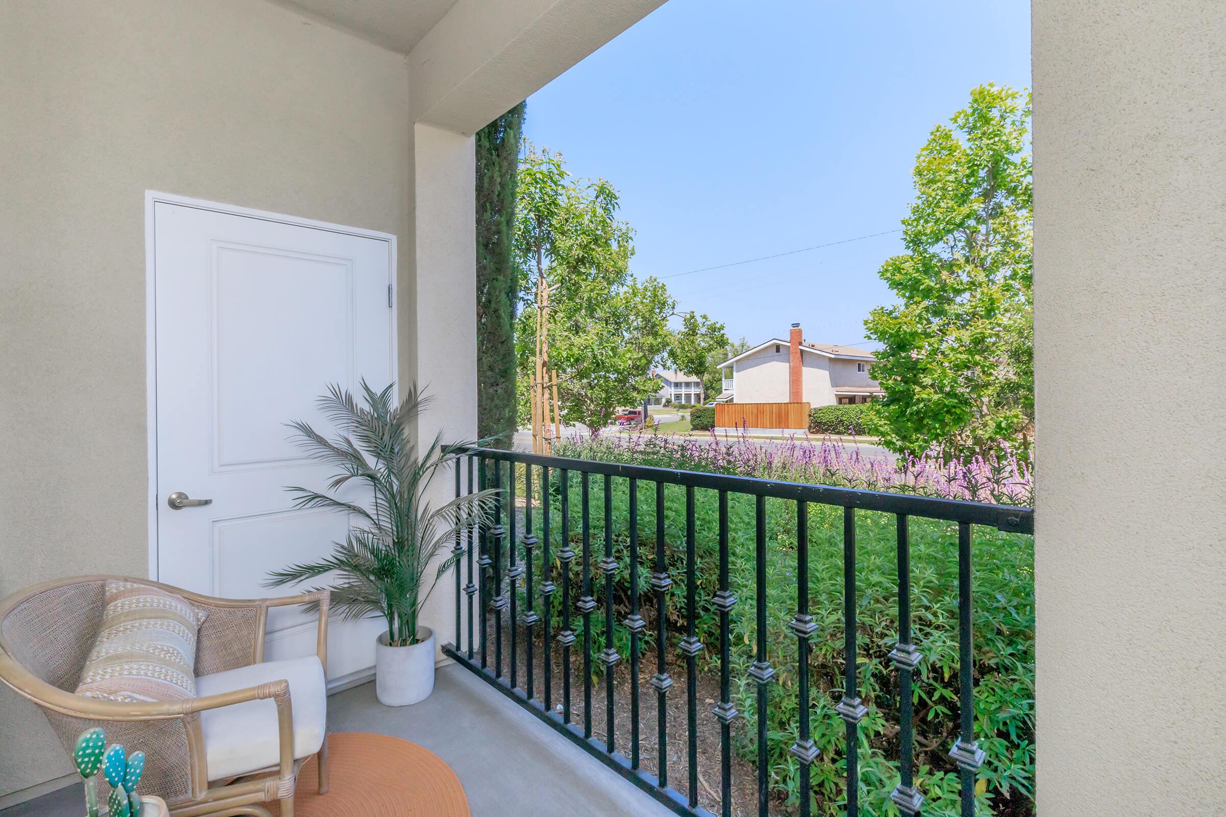 A view from a balcony featuring a wicker chair with a patterned cushion, a small potted plant, and a round outdoor rug. Greenery and flowering plants are visible below the balcony railing, with houses and trees in the background under a clear blue sky.