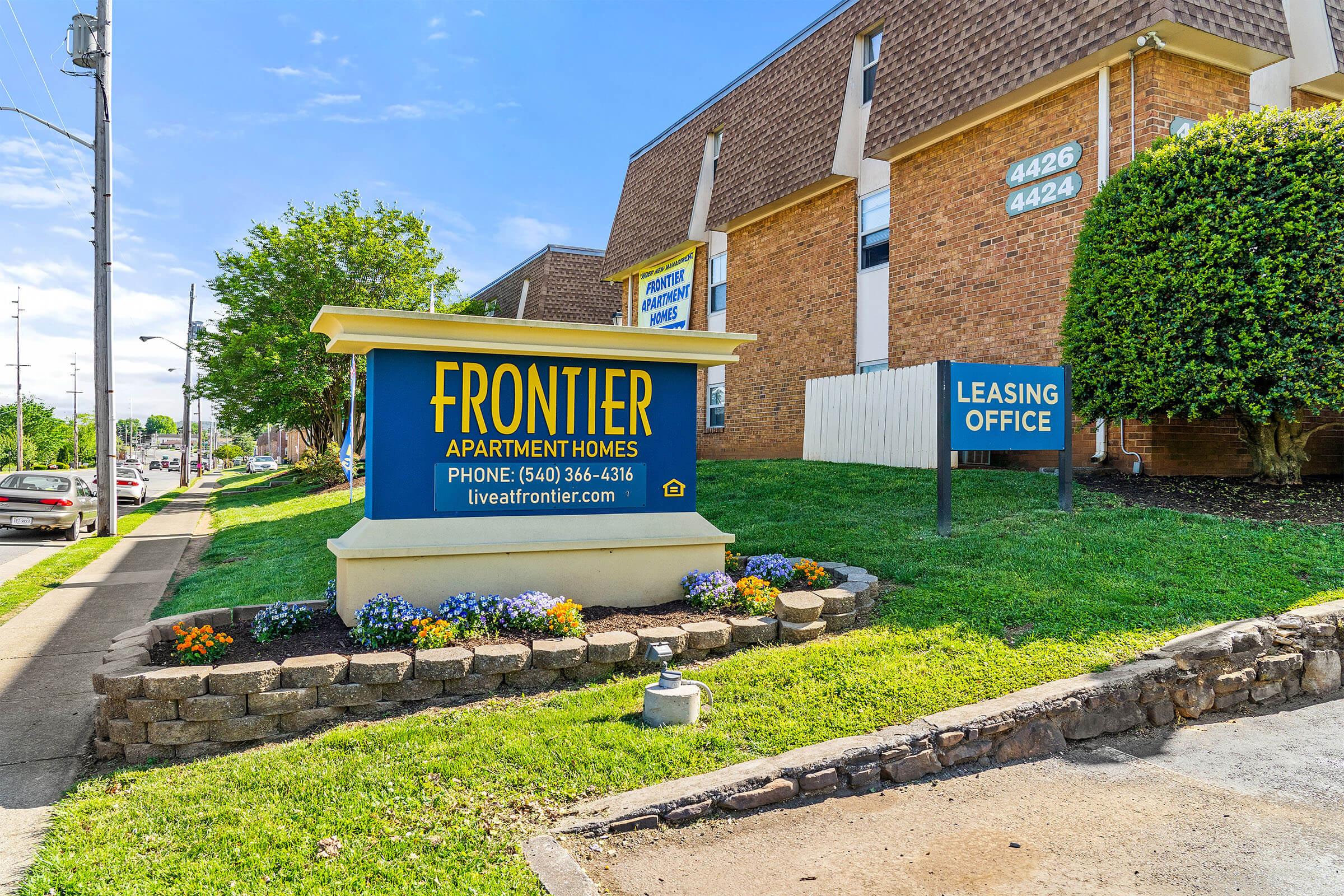 Sign for Frontier Apartment Homes featuring the leasing office. The sign is prominently displayed near the building, surrounded by colorful flowers and greenery. The address numbers “4426” and “4442” are visible on the building. It’s a bright, sunny day with clear blue skies.