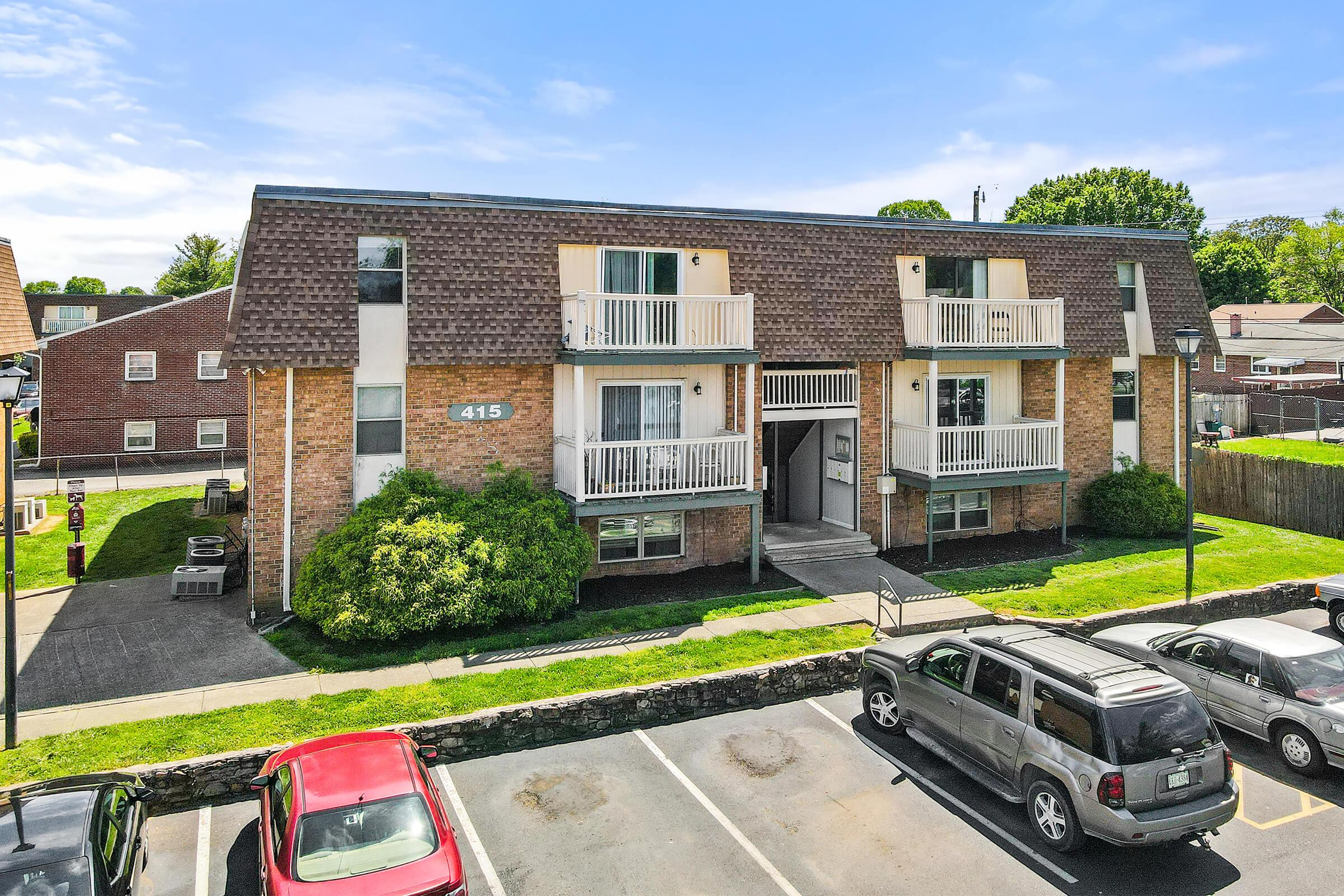 A two-story brick apartment building with white balconies, marked with the number 415. Lush greenery surrounds the building, and a parking lot with several cars is visible in the foreground. The sky is clear and blue, creating a bright and welcoming atmosphere.