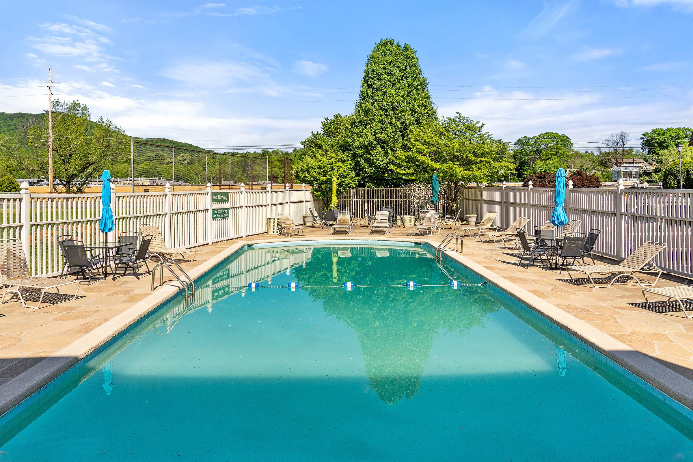 A clear, inviting swimming pool surrounded by lounge chairs and umbrellas, set in a landscaped area with trees in the background. The scene reflects a sunny day with blue skies, providing a tranquil atmosphere for relaxation and leisure.