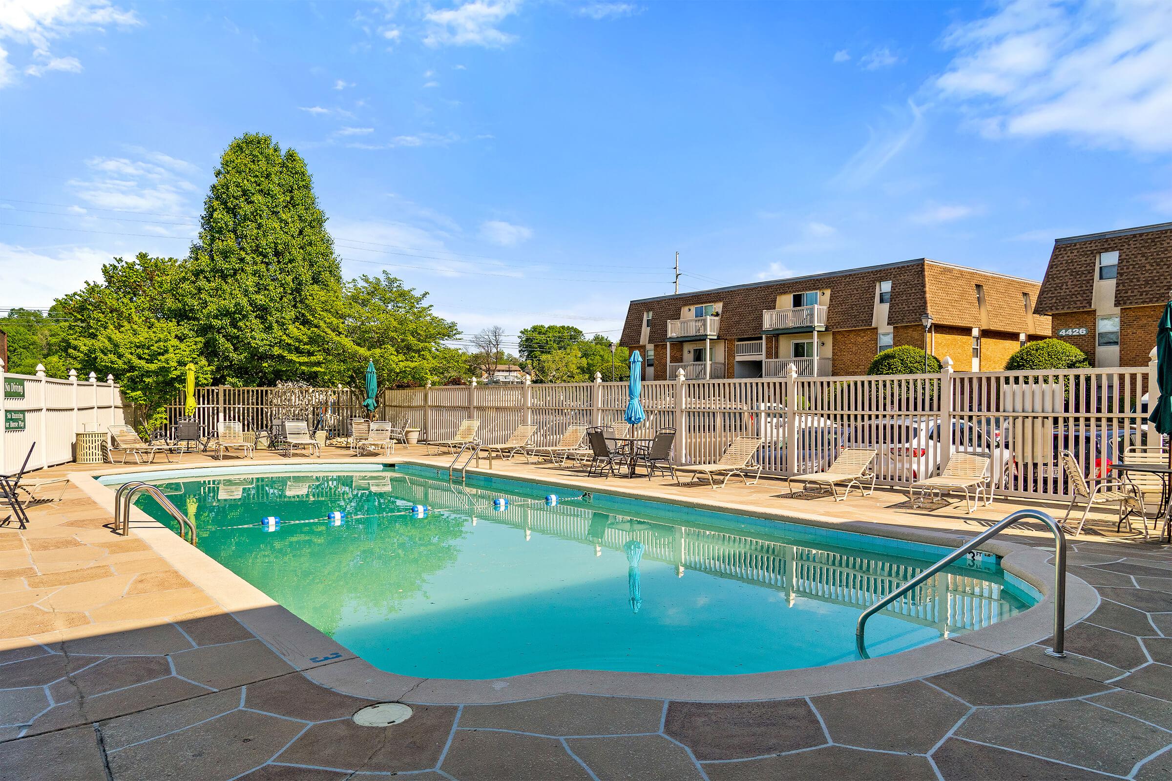A clear blue swimming pool bordered by lounge chairs and umbrellas, set in a clean, well-maintained outdoor area. Surrounding greenery and a brick building provide a pleasant backdrop, all under a bright blue sky with a few clouds.