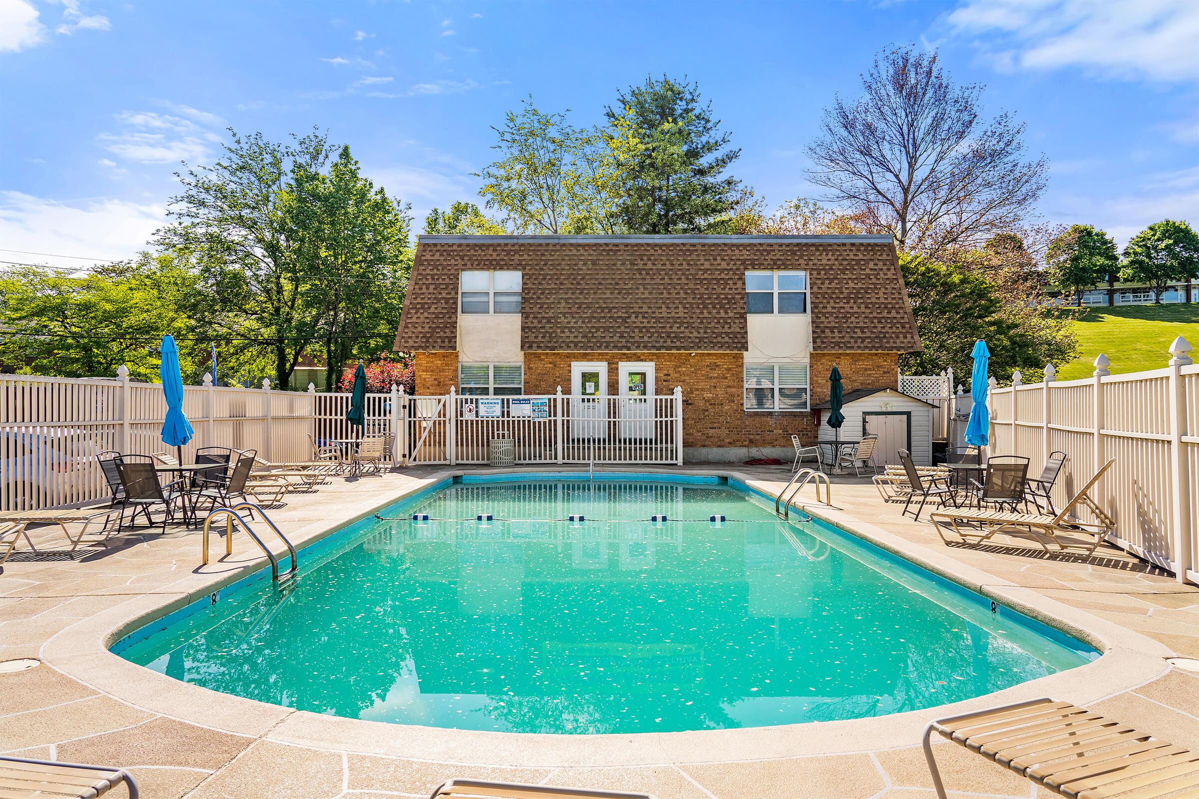 A clear blue swimming pool surrounded by a fenced area with lounge chairs and umbrellas. A two-story brick building is visible in the background, with trees and greenery nearby. The sky is bright and sunny, indicating a pleasant outdoor setting.