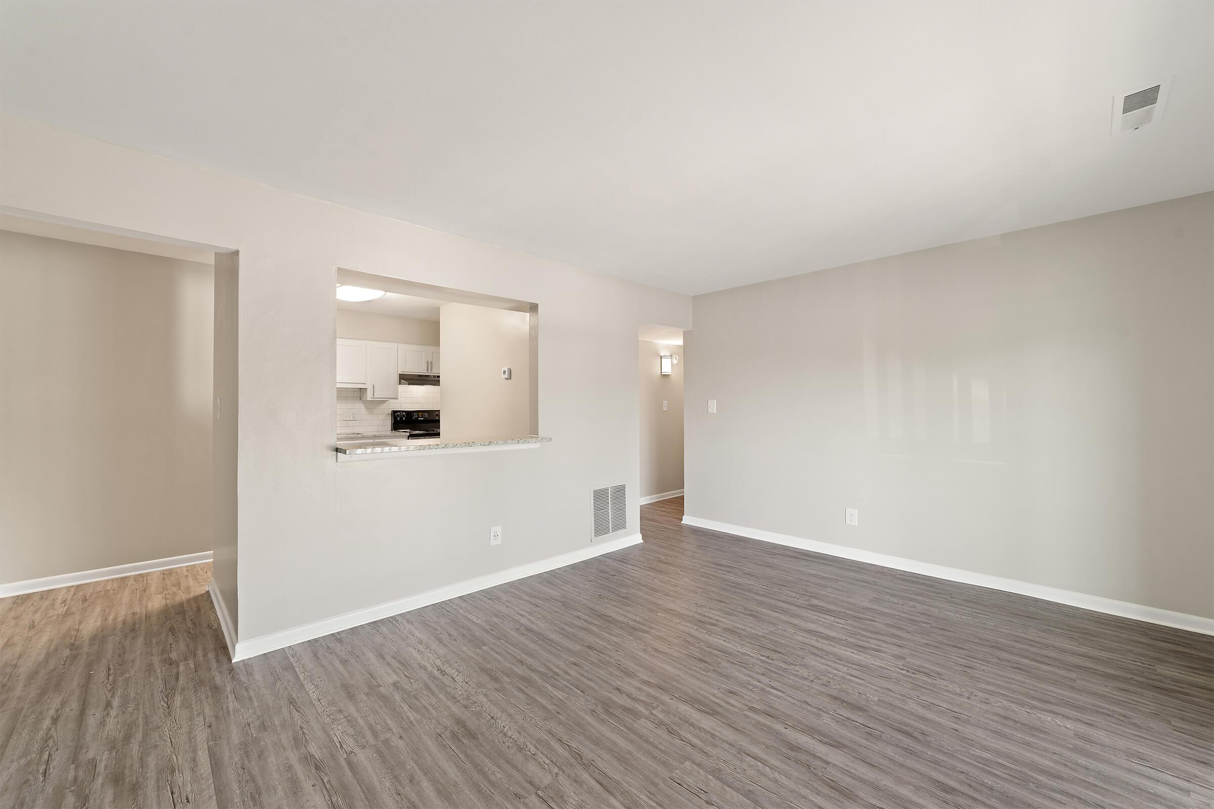 A spacious, empty living room with light grey walls and vinyl flooring. A doorway leads to a kitchen partially visible through an open wall. The room has ample natural light and modern fixtures, creating a bright and inviting atmosphere.