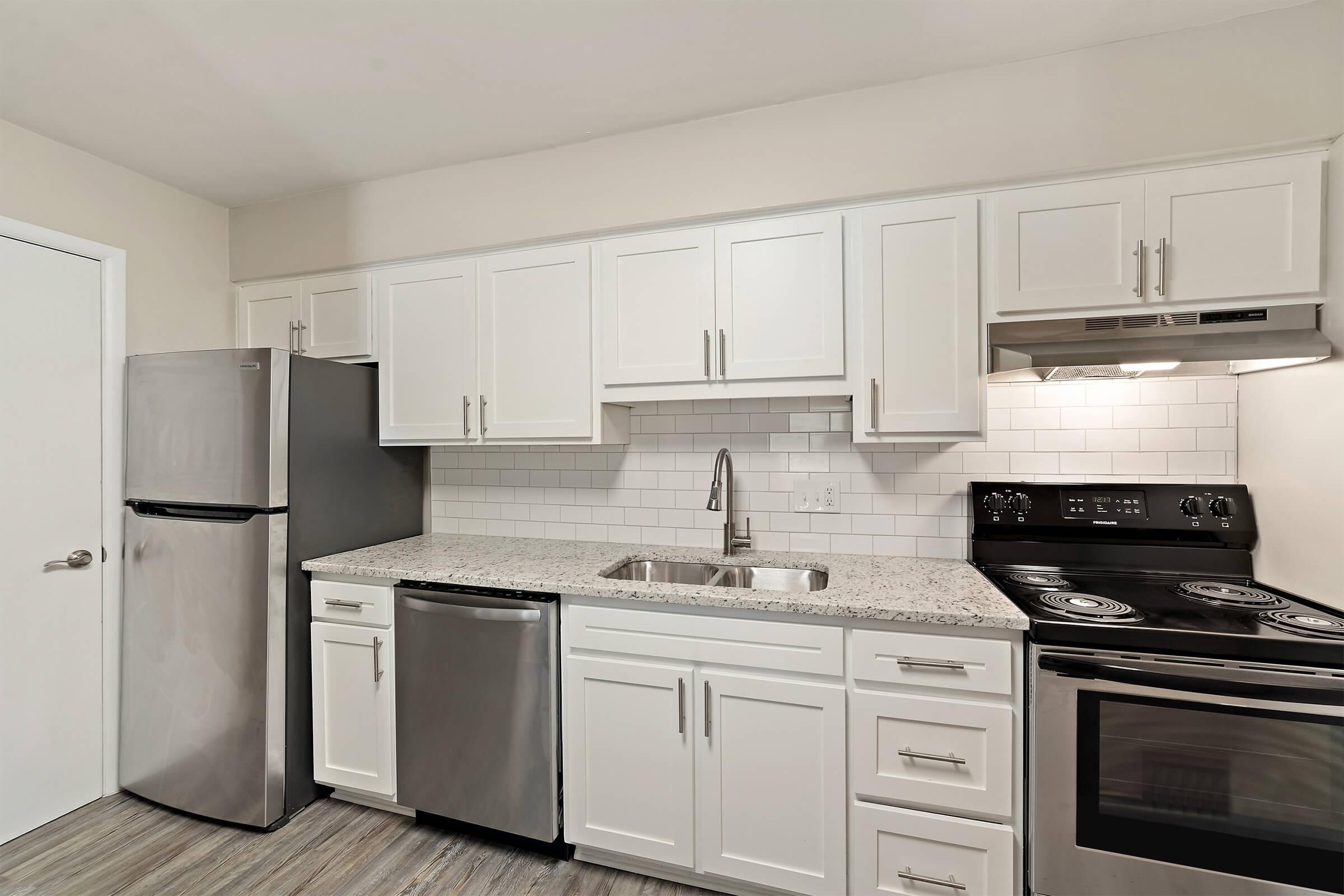 Modern kitchen featuring white cabinetry, a stainless steel refrigerator and dishwasher, and a black stove. The countertop is made of granite with a subtle pattern, and the backsplash is white subway tile. Soft lighting illuminates the space, creating a clean and inviting atmosphere.